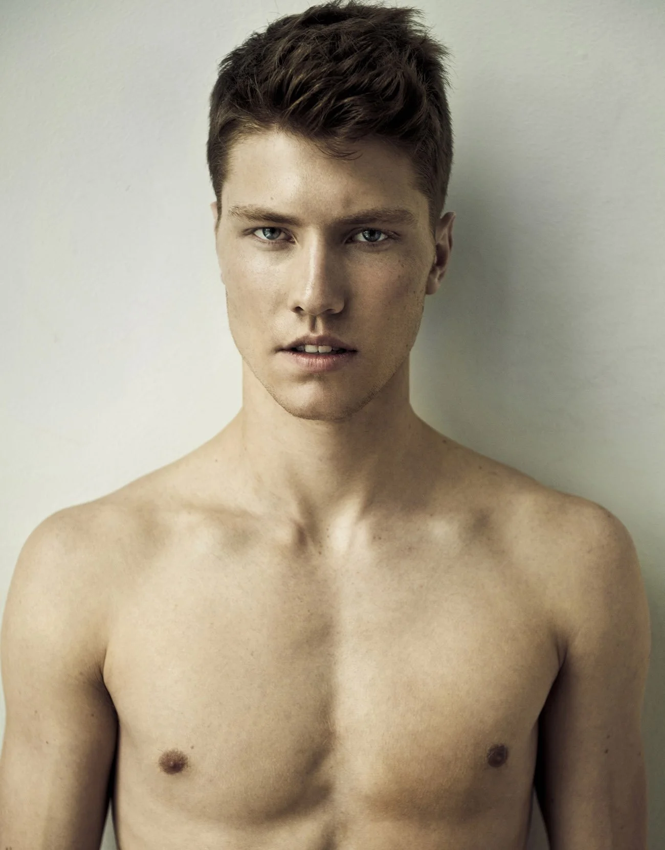 A young man with short brown hair and blue eyes, shirtless, standing against a plain off-white wall.