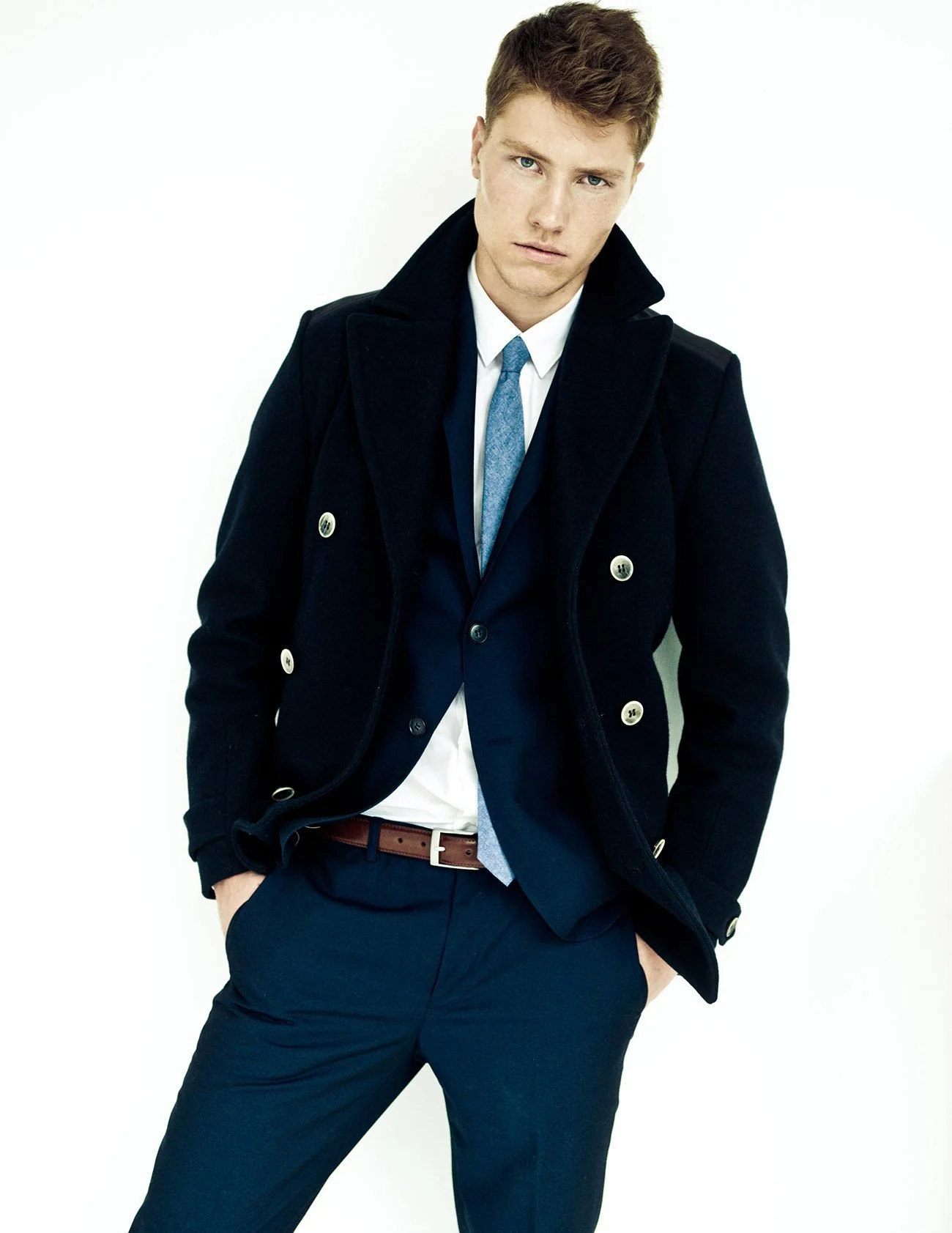 A young man with brown hair and blue eyes wearing a dark navy peacoat, white shirt, blue tie, navy pants, and a brown belt, standing against a plain white background.
