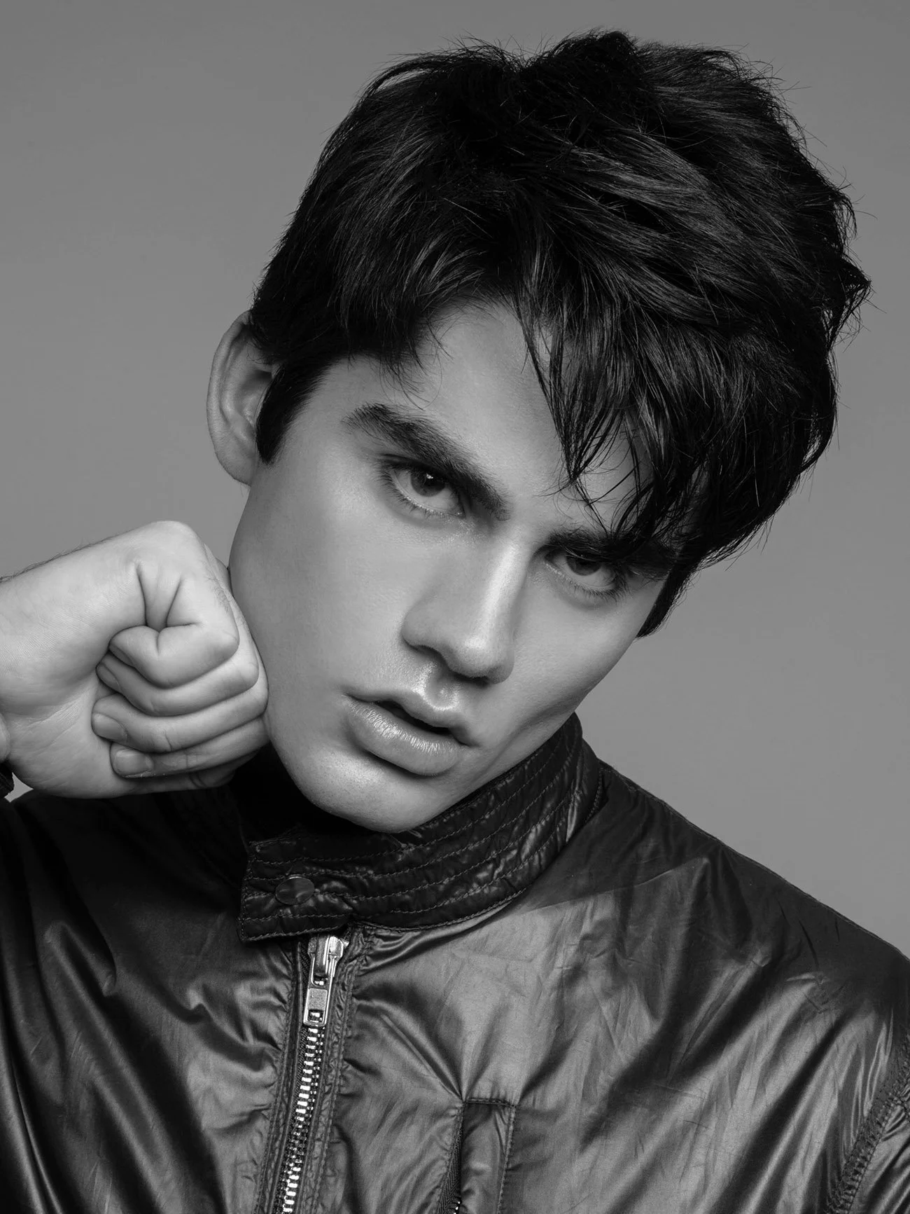 Black and white portrait of a young man with tousled dark hair, wearing a shiny black leather jacket, looking intensely into the camera with his hand near his face.