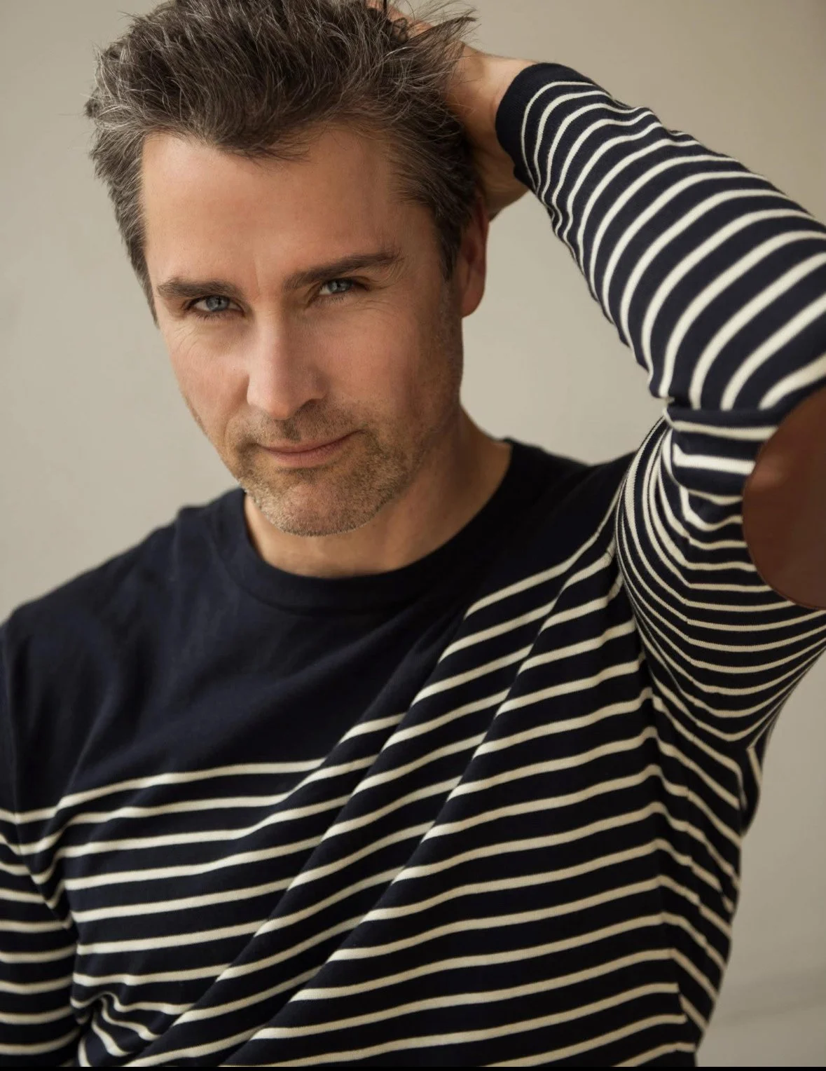 A man with light skin, gray hair, and blue eyes, wearing a black shirt and a black and white striped long sleeve shirt, posing with one hand behind his head.
