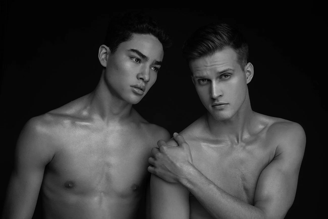 Black and white photo of two shirtless men with serious expressions, standing close together against a dark background.