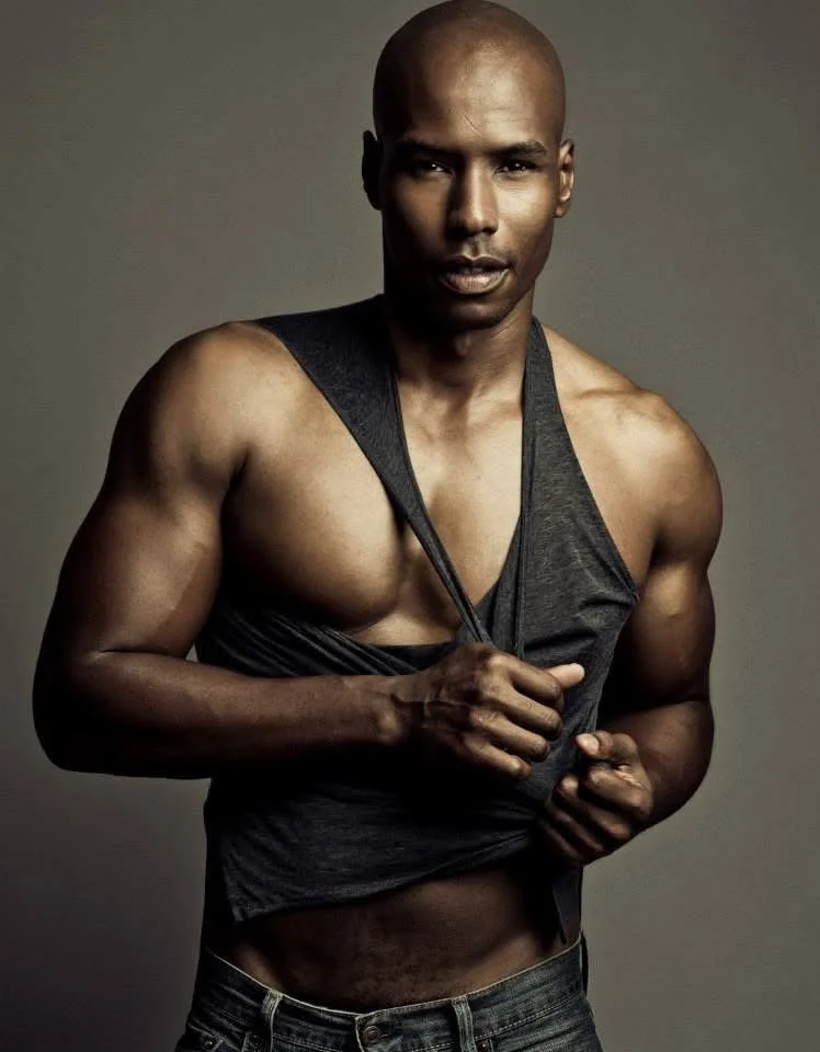 A muscular man with dark skin and a shaved head, wearing a sleeveless dark shirt and jeans, holding the shirt with one hand and looking at the camera against a plain background.