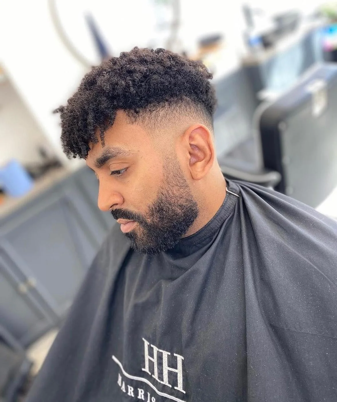 A man with curly hair and a beard sitting in a barber's chair, wearing a black barber cape, at a barbershop.