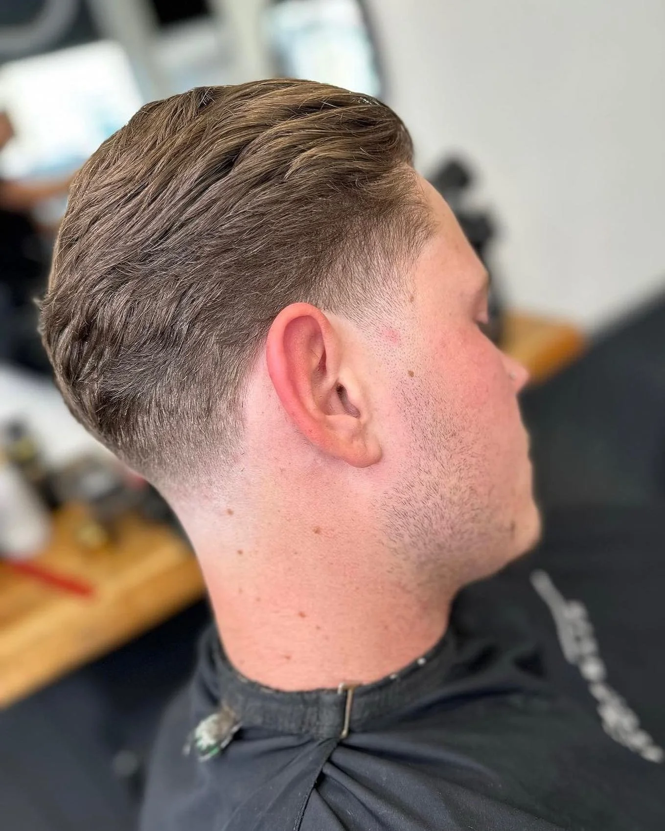 Side view of a young man with a classic undercut hairstyle, with his hair on top combed back and short faded sides, sitting in a barbershop chair.