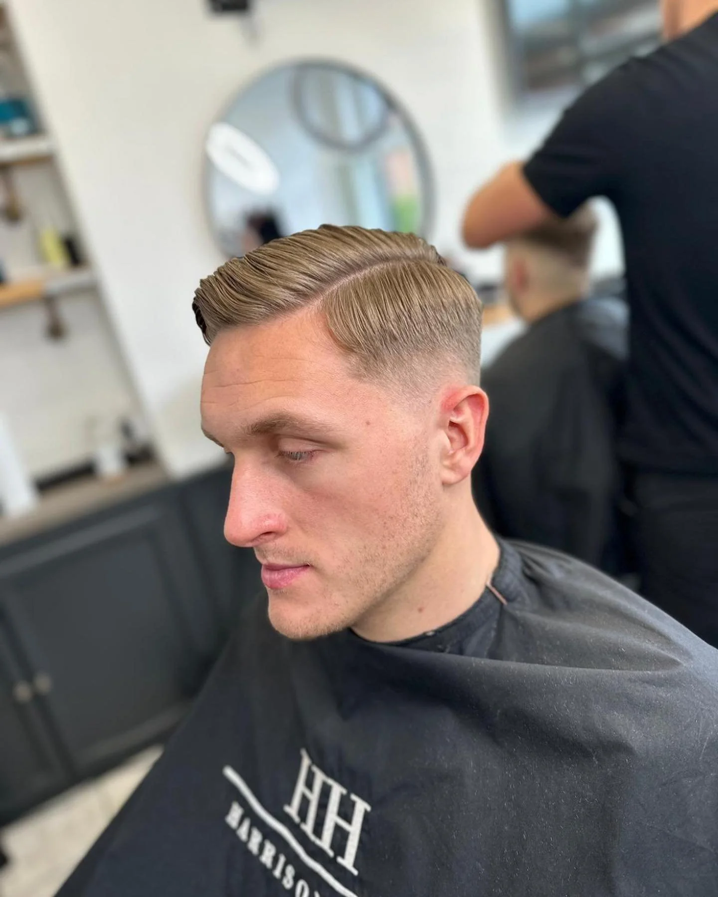 Young man with a fresh hairstyle featuring a side part and slicked back top, sitting in a barber chair at a barbershop.