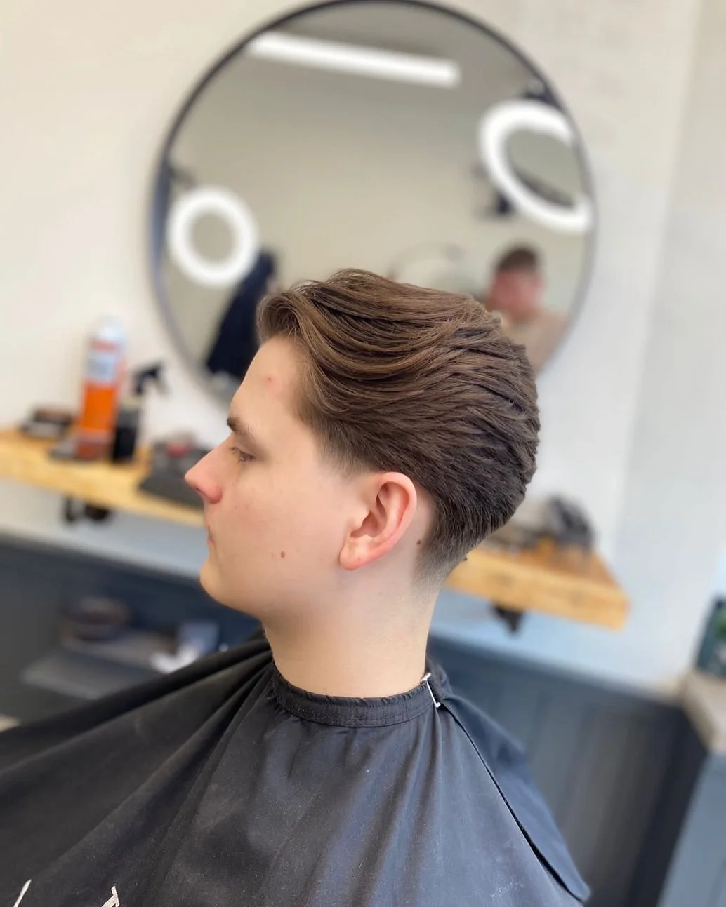 Young man with styled brown hair sitting in a barber's chair, reflected in a round mirror in a barbershop.