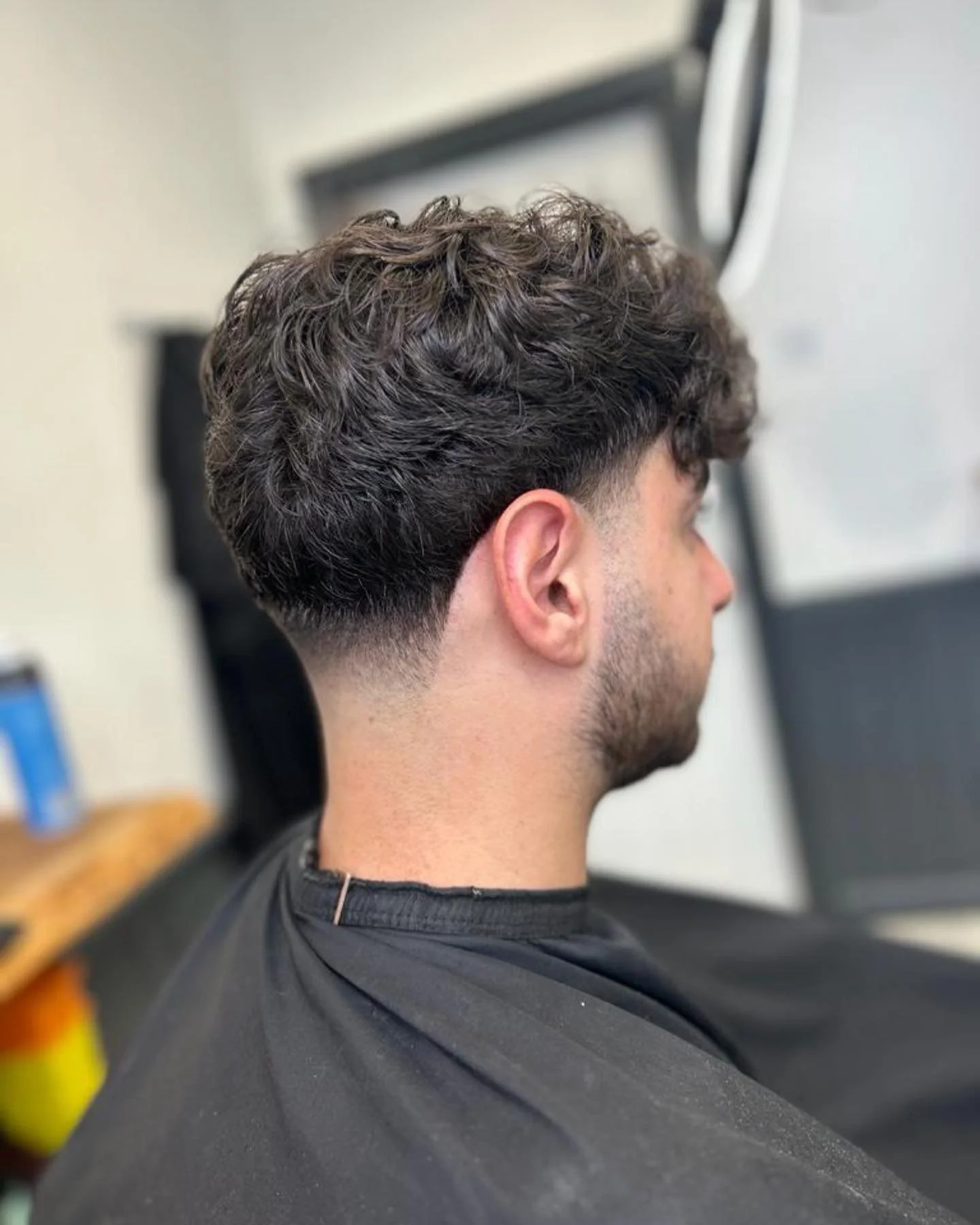 Side view of a young man with dark, curly hair in a barber's chair, showing a fresh fade haircut with longer curls on top.