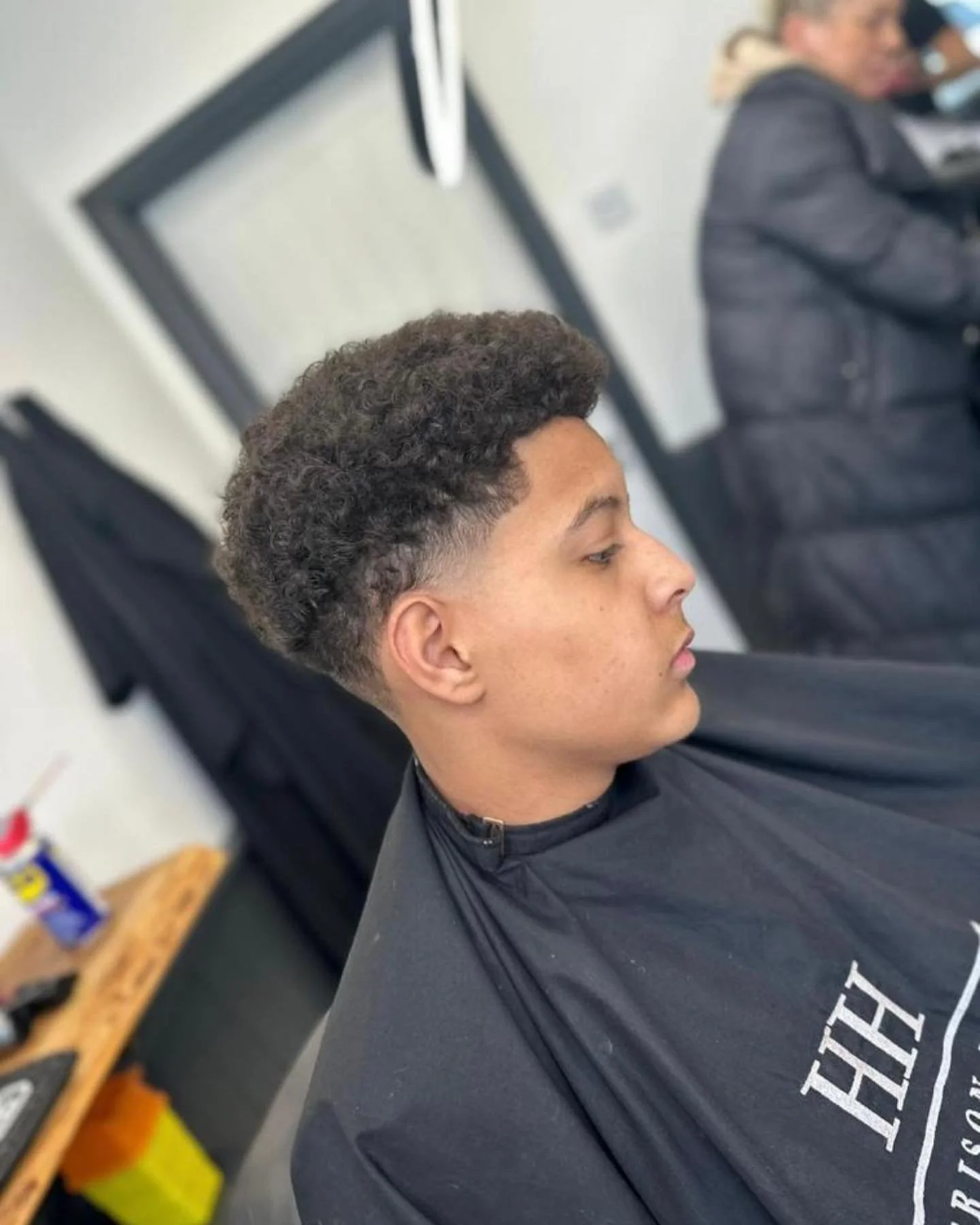 Young man with curly, styled hair sitting in a barber's chair at a haircutting salon.
