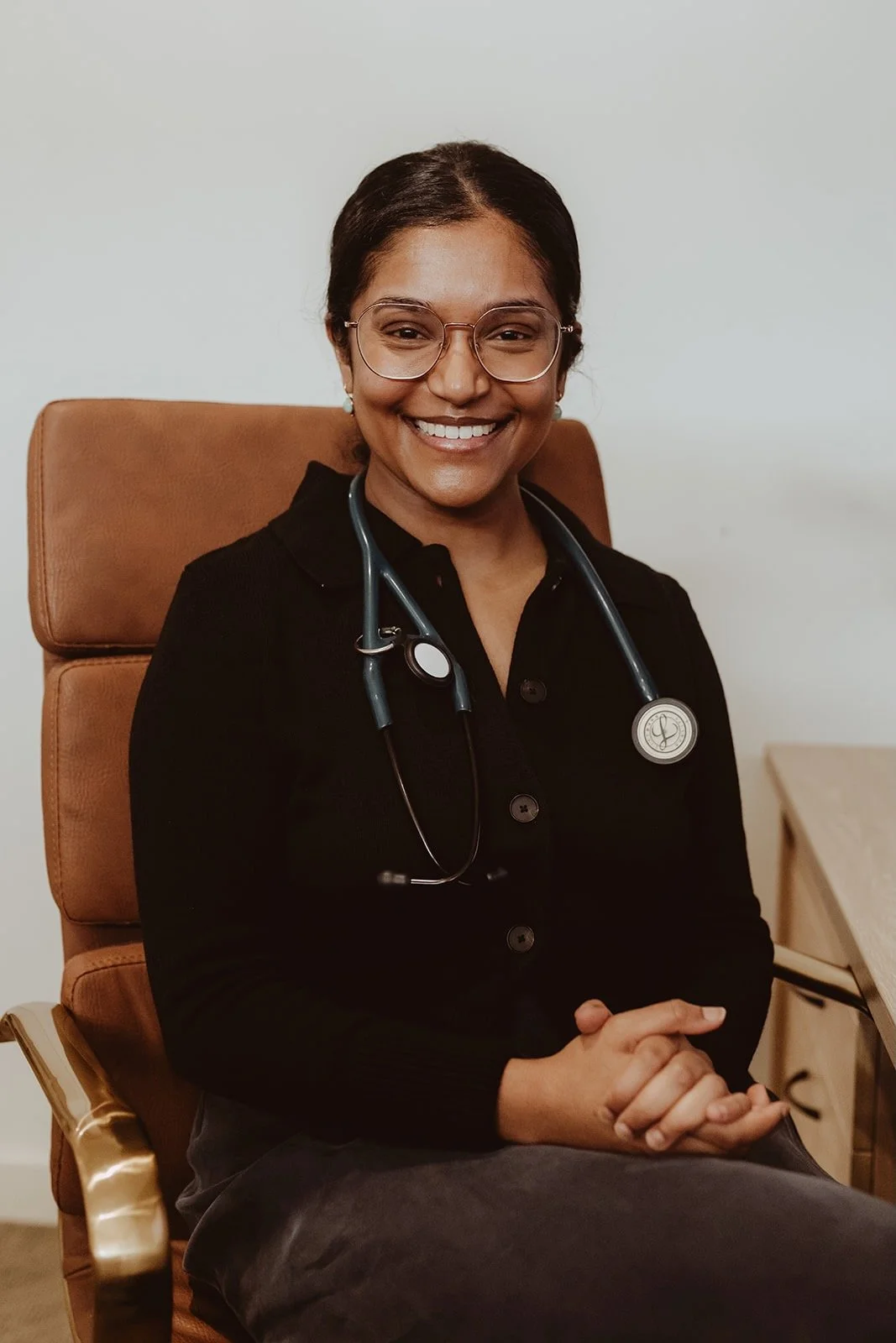 A smiling female healthcare professional sitting in a tan leather chair, wearing glasses, a black top, and a stethoscope around her neck.