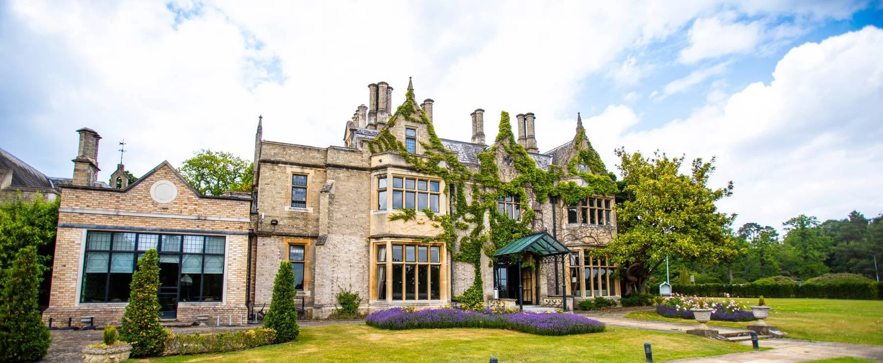 Historic stone mansion with ivy-covered walls, large windows, and a landscaped garden with purple flowers and a tree, under a partly cloudy sky.