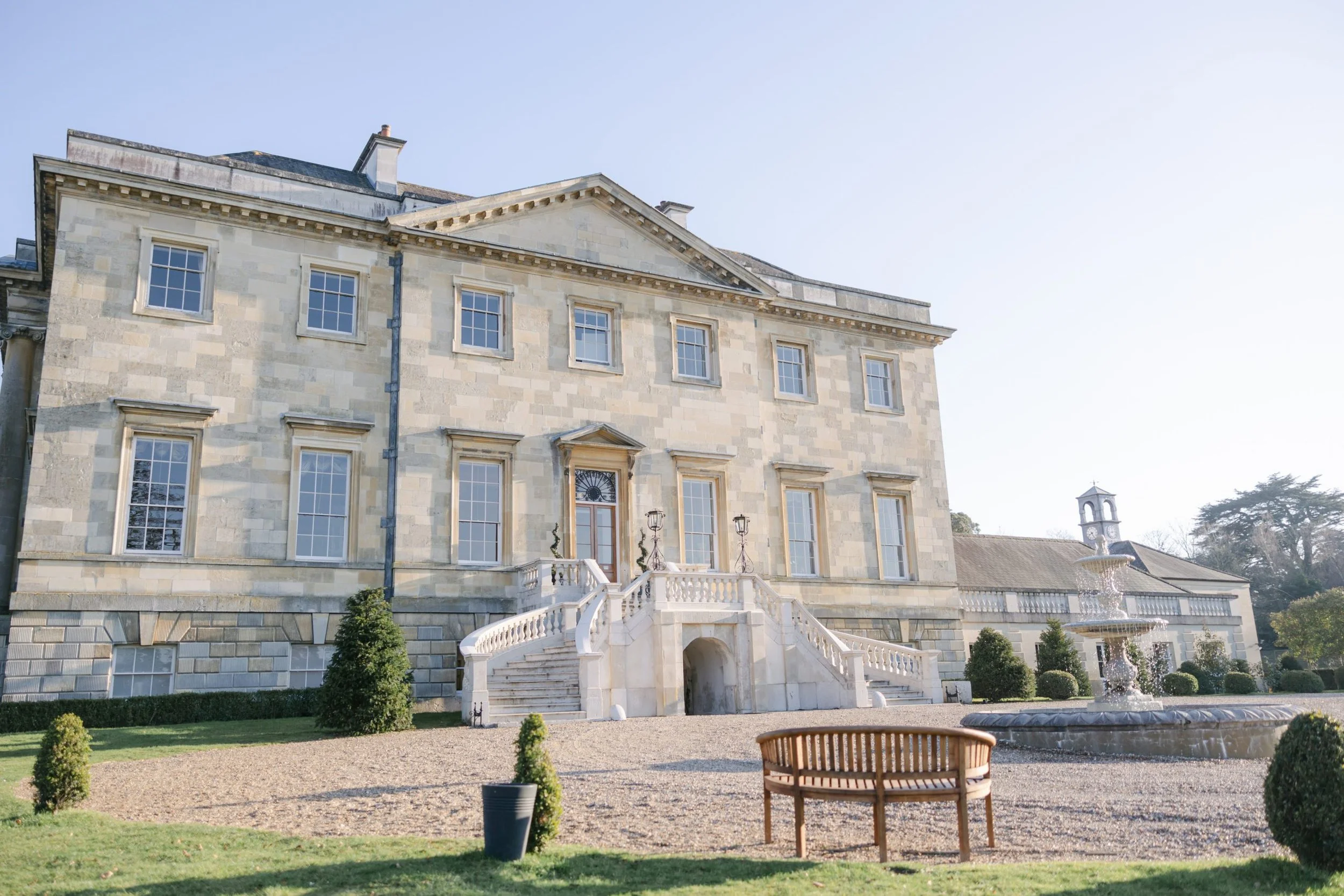Large historic mansion with stone facade, multiple tall windows, grand staircase, landscaped lawn, fountain, and bench in the foreground.
