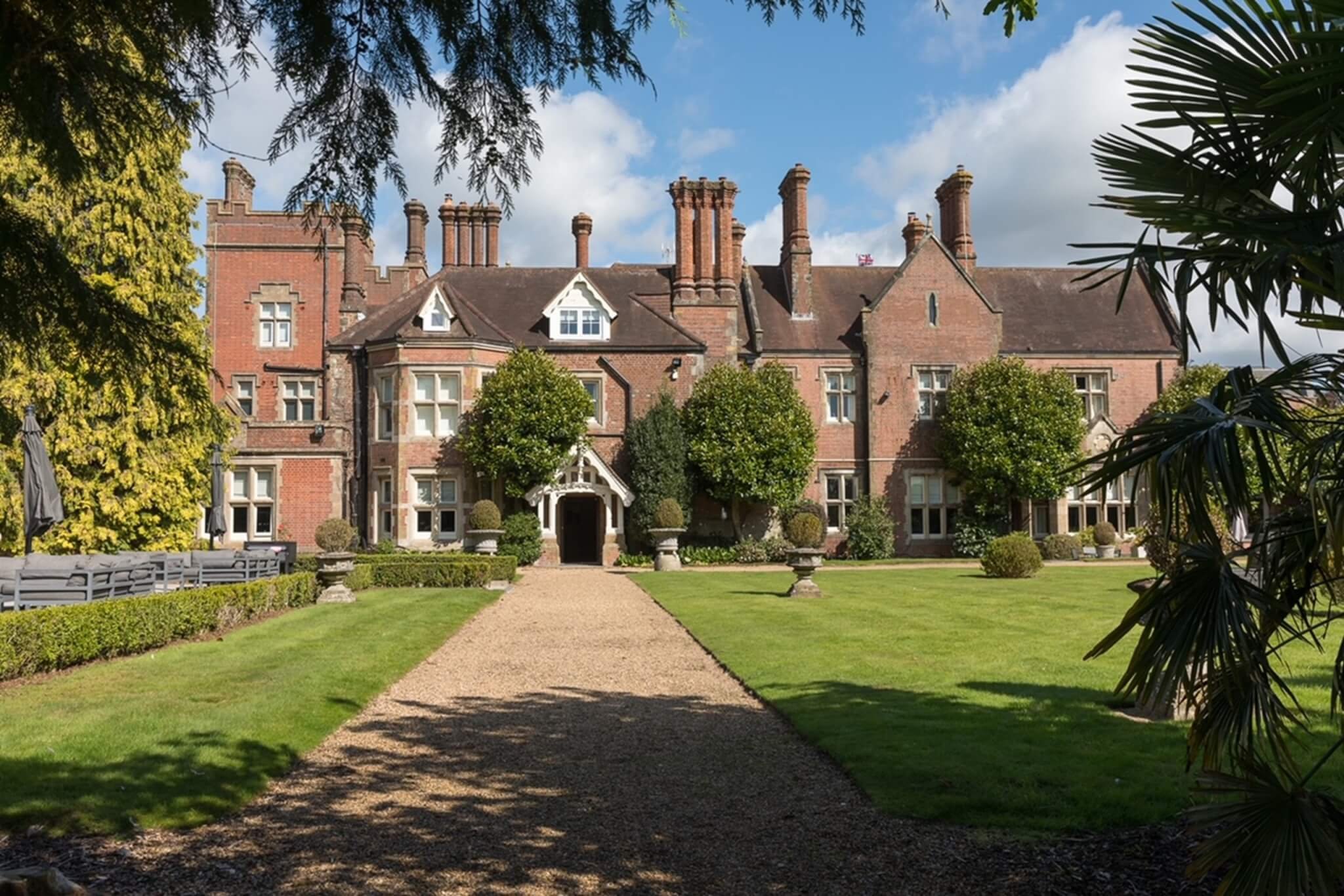 Large historic brick mansion with multiple chimneys, surrounded by a well-maintained garden with trimmed bushes, trees, and a central gravel pathway leading to the entrance, under a partly cloudy sky.