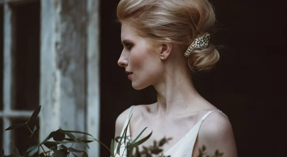 Profile of a woman with elegant updo hairstyle, wearing pearl earrings and a white dress, standing outdoors near a wooden structure with greenery.