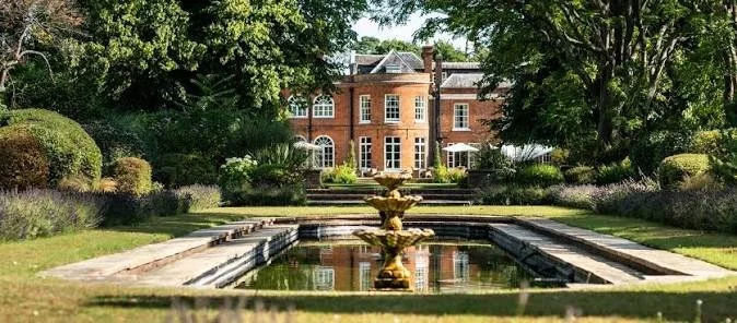 Large historic brick mansion with multiple chimneys, surrounded by a well-maintained garden with trimmed bushes, trees, and a central gravel pathway leading to the entrance, under a partly cloudy sky.