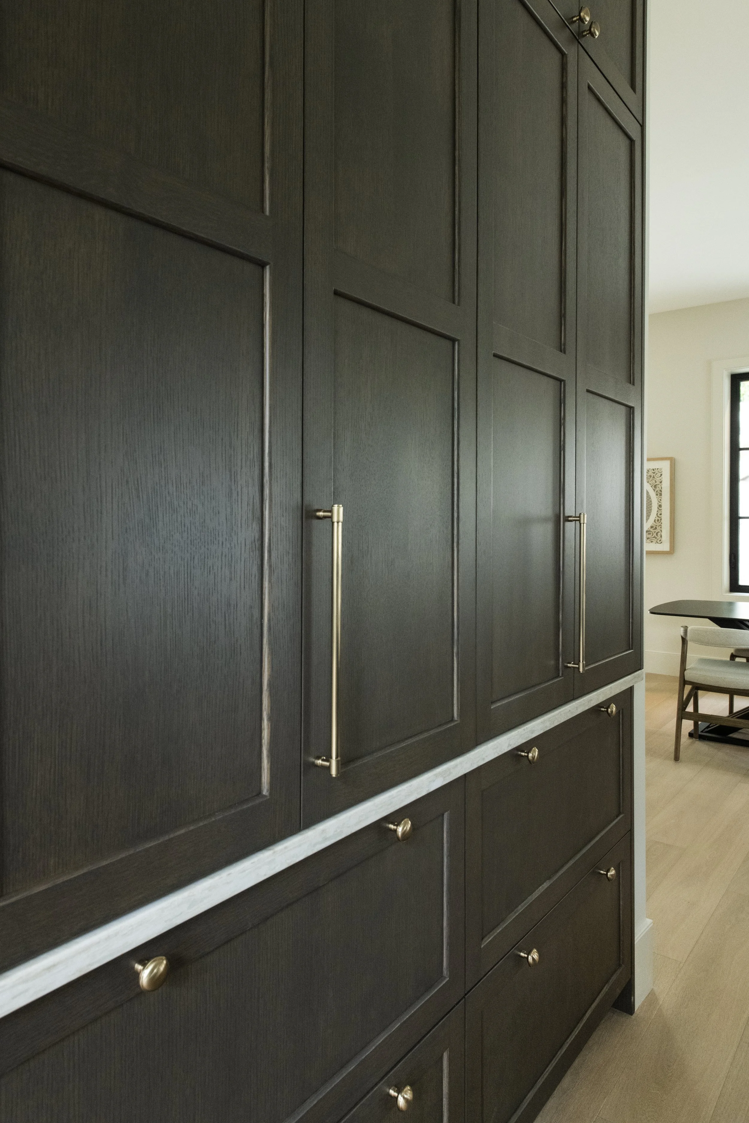 Close-up of tall, dark wooden cabinets with gold handles in a modern kitchen or dining area, with a table, chairs, and a window visible in the background.