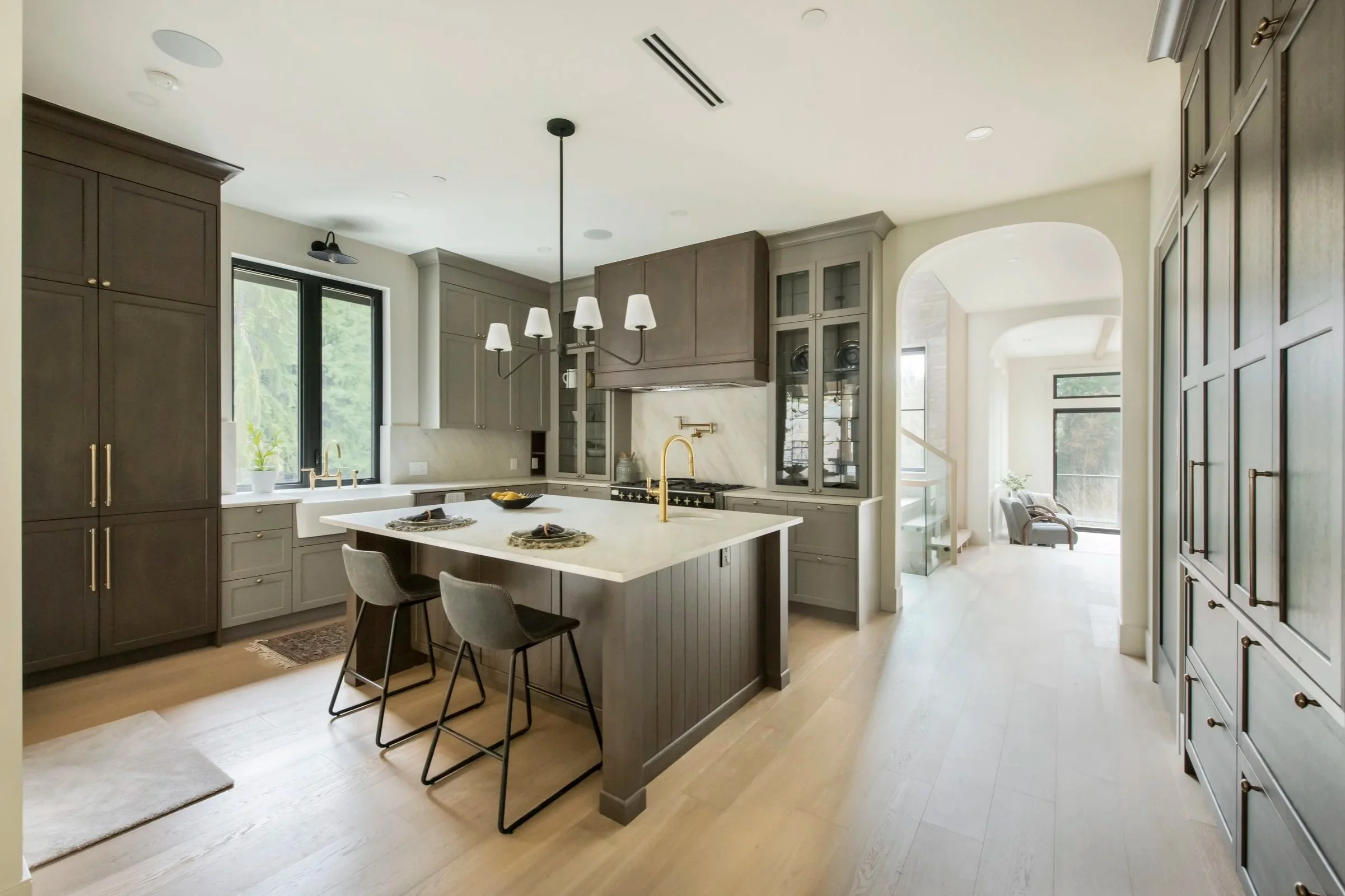 Modern kitchen with gray cabinetry, white island with gold faucet, black barstools, and large window with greenery outside.