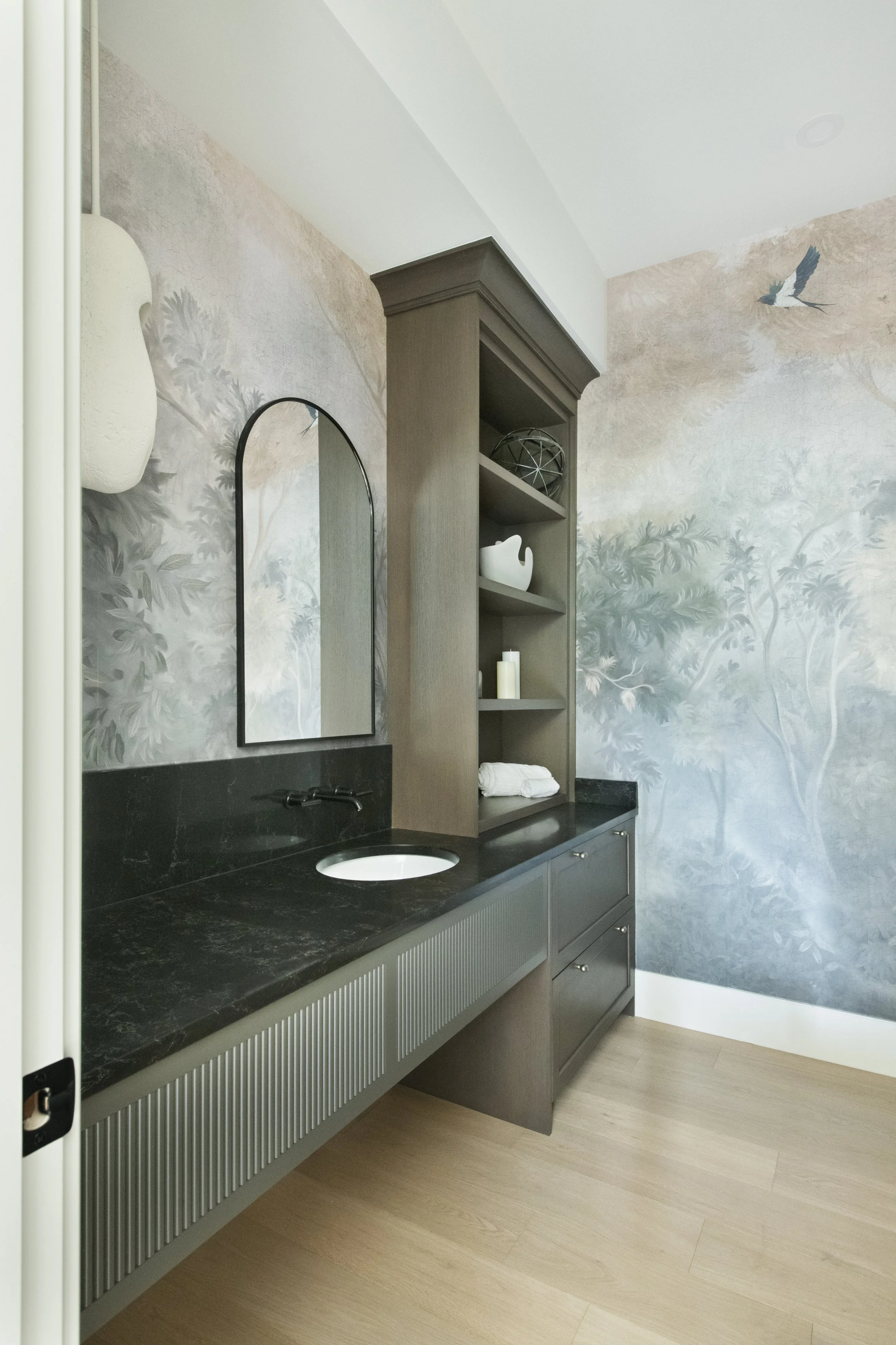 Modern bathroom with a black marble countertop, built-in sink, tall wooden shelving unit, mirror, and nature-themed wall mural featuring trees and a flying bird.