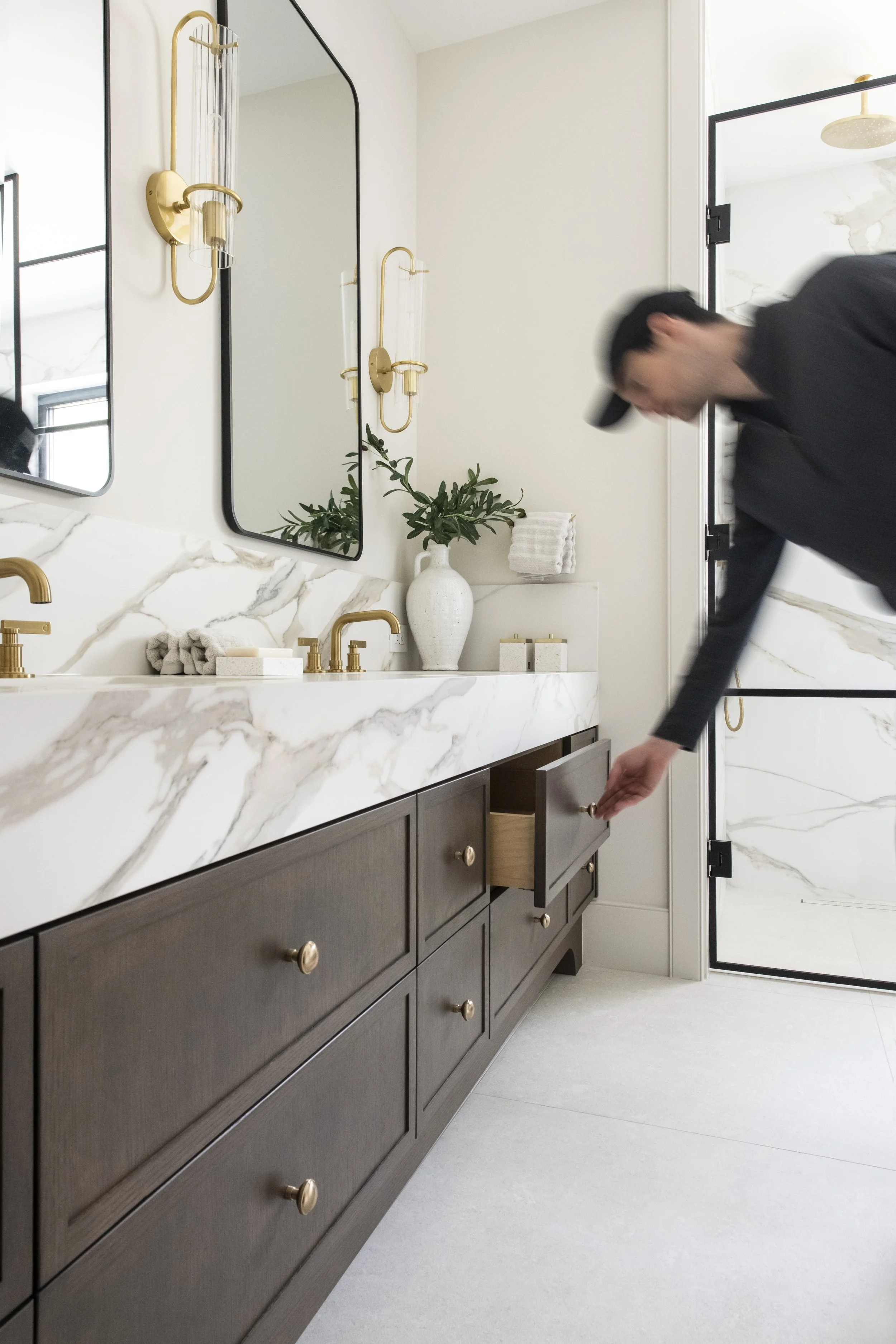 A person opening a drawer beneath a marble bathroom countertop with gold fixtures, framed mirrors, a white vase with greenery, and rolled towels on a shelf.