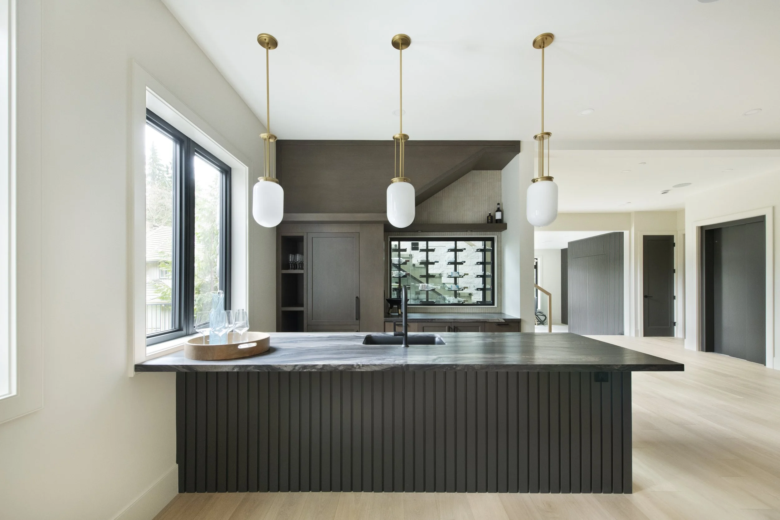 Modern kitchen with black island, pendant lights, and large window, open to living space with stairs and cabinetry.