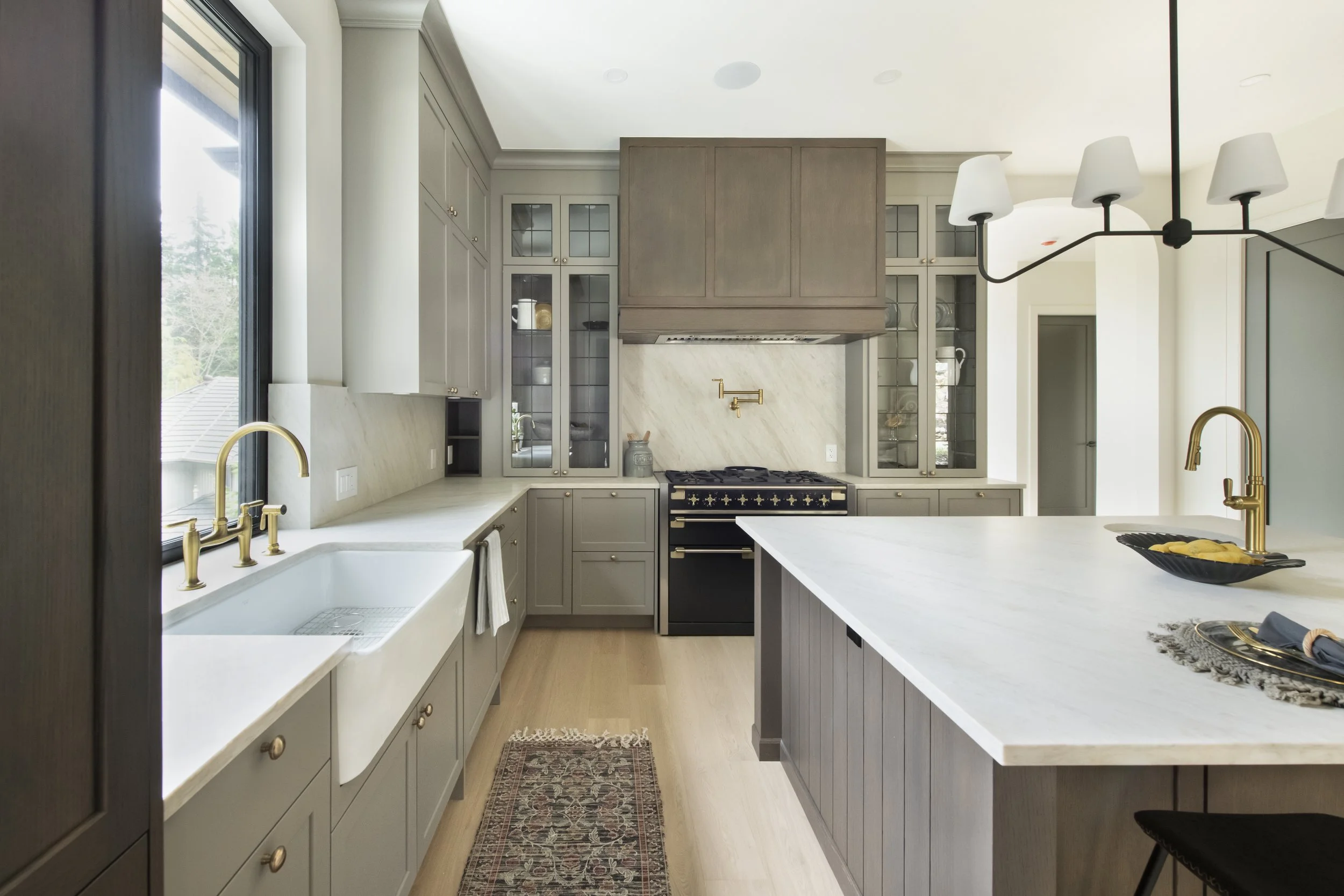 Modern kitchen with light gray cabinets, white countertops, black and gold stove, farmhouse sink, and a kitchen island with a white marble top.