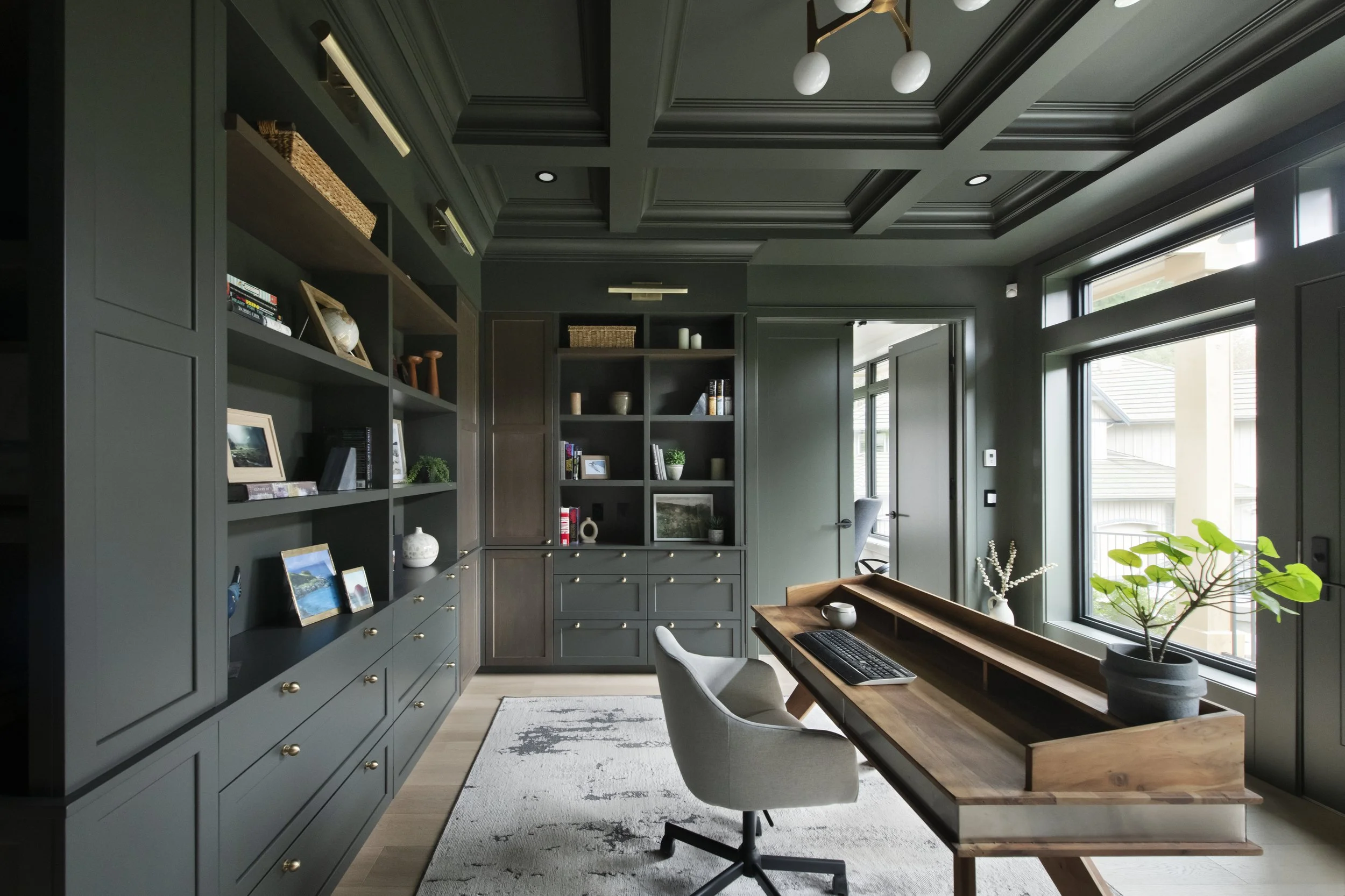 Modern home office with dark green built-in bookshelves, a wooden desk with a computer, a gray office chair, large windows, and decorative items such as framed photos, plants, and vases.