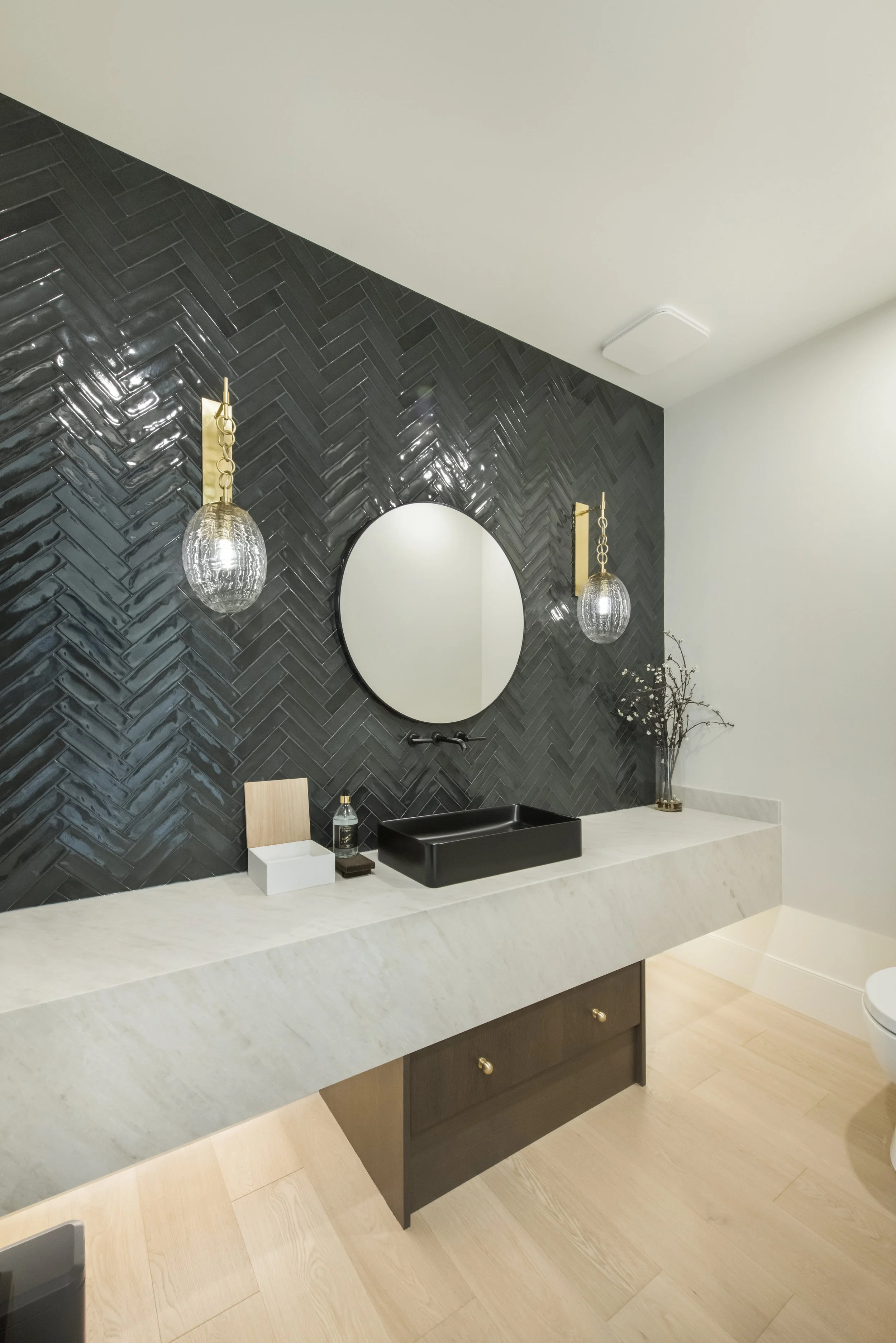 Modern bathroom with black textured tile wall, round mirror, gold pendant lights, white marble countertop, black vessel sink, and decorative vase with branches.