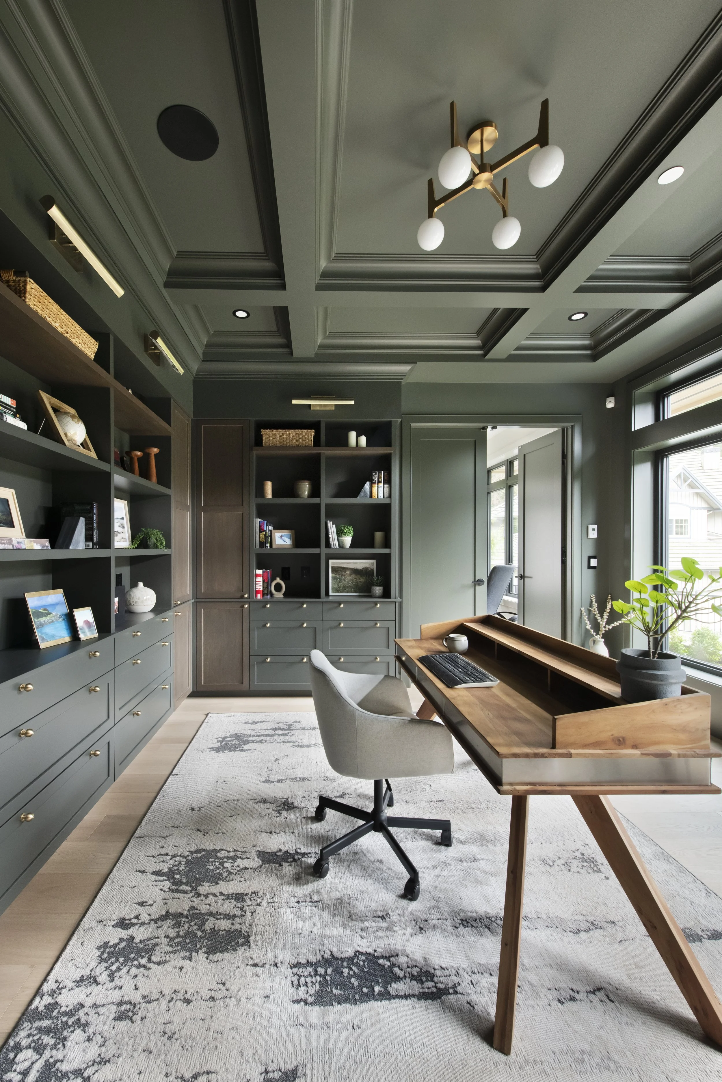 Modern home office with dark green walls, built-in shelves, a wooden desk, an office chair, and large windows letting in natural light.