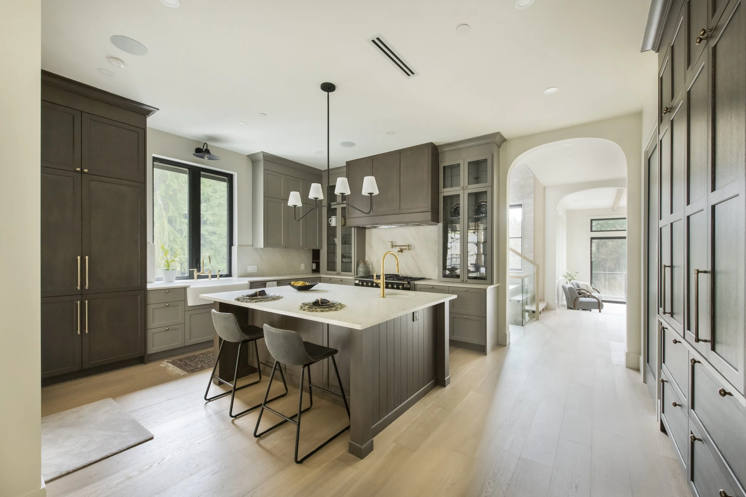 Modern kitchen with gray cabinetry, white island with gold faucet, black chairs, and large windows.
