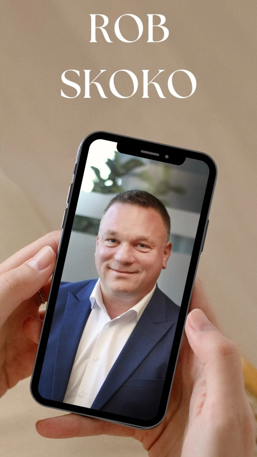 A person holds a smartphone displaying a picture of a man in a blue suit and white shirt. The background is blurred with some plants. The text above reads 'Rob Skoko' in large white letters.