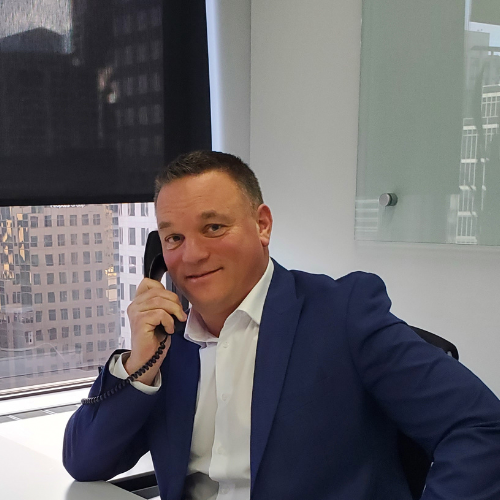 A man in a white shirt and blue blazer sitting at a desk, speaking on a landline phone, with a cityscape visible through a window behind him.