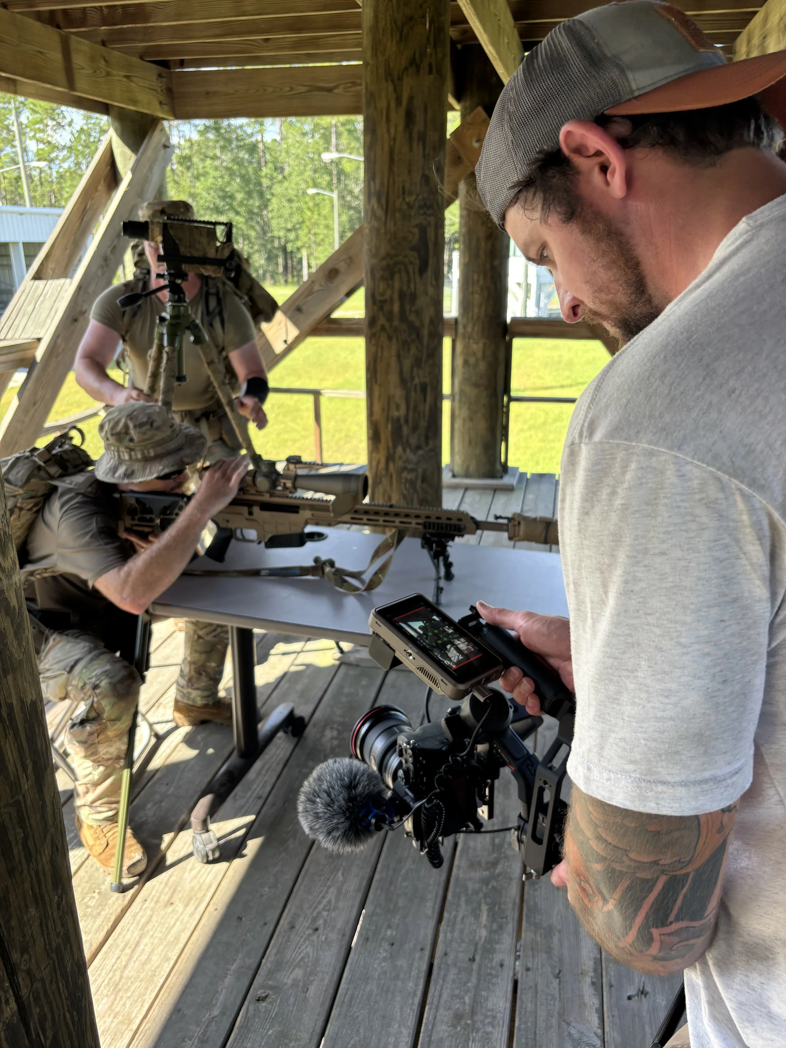 A man on the right holding a camera with a microphone, filming two men in military gear aiming a sniper rifle, inside a wooden structure with natural light and trees outside.