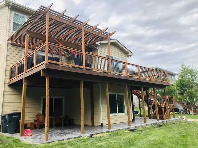 A large two-story house with a newly built wooden deck on the upper level and stairs leading down to the yard. The deck has a pergola on top with slats, and the house has beige siding and white-framed windows.