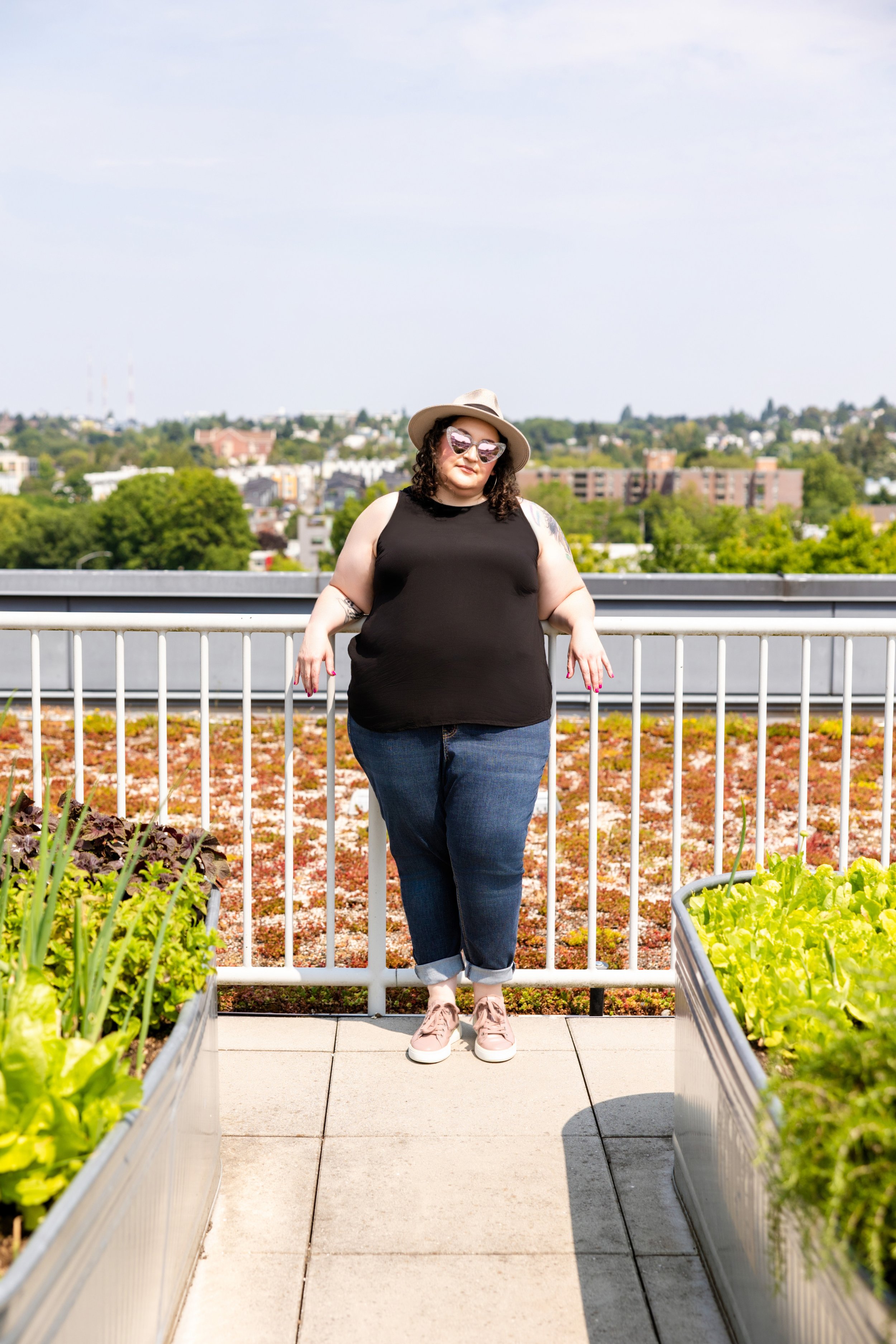 Golden Poppy Digital Owner Tori standing outside leaning on a railing. She is wearing a hat, sunglasses, a black shirt, jeans, and sneakers.