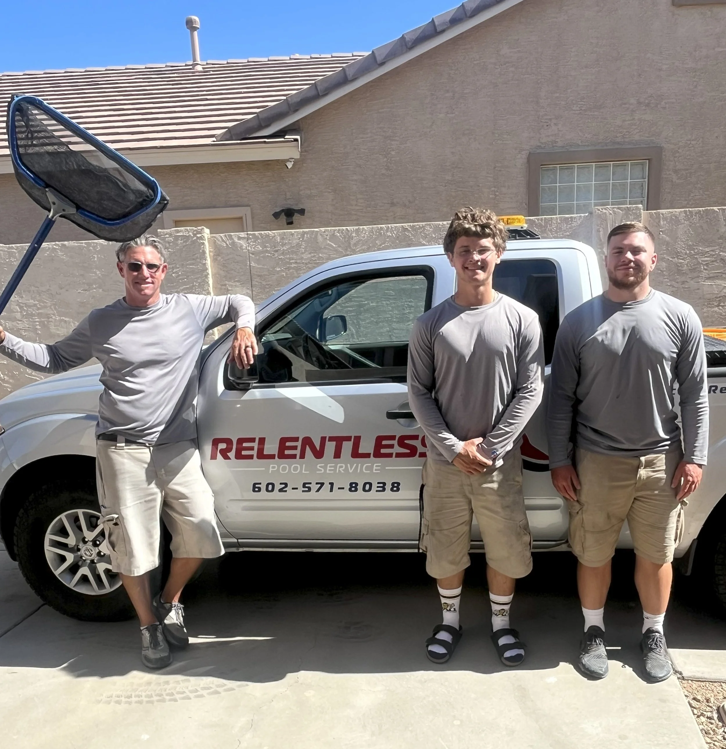 Three men standing and leaning against a white pickup truck with the words "Relentless Pool Service" and a phone number on the side. The man on the left is holding a pool skimmer. All three are wearing gray shirts and shorts, standing outdoors on a sunny day, with a house and blue sky in the background.
