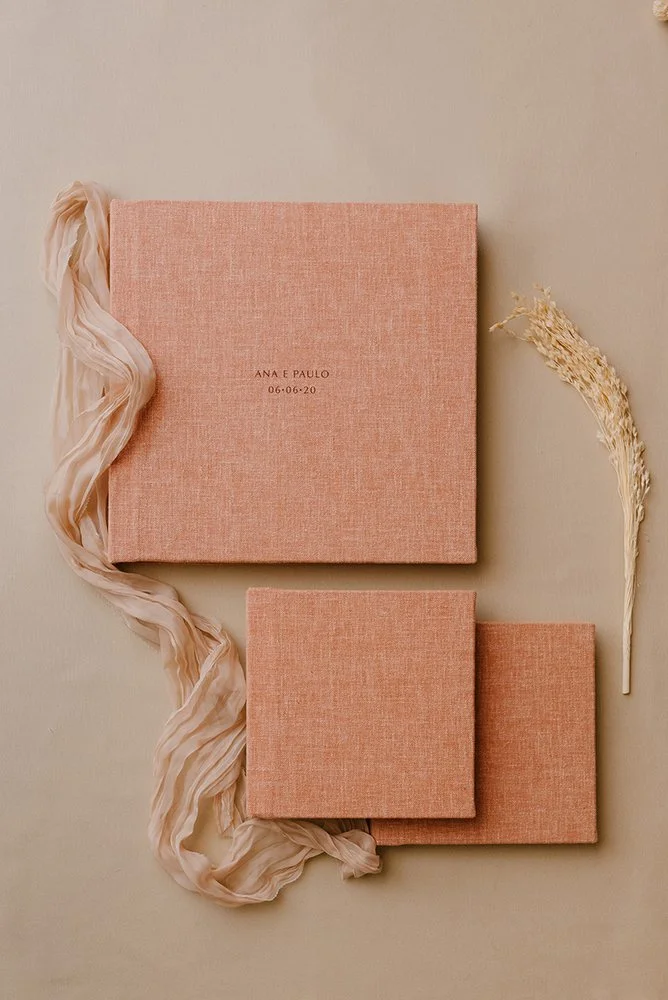 Álbum de fotos com capa cor salmão, com texto personalizado e data, acompanhado de tecido de seda e uma espiga de trigo seca, sobre fundo claro.