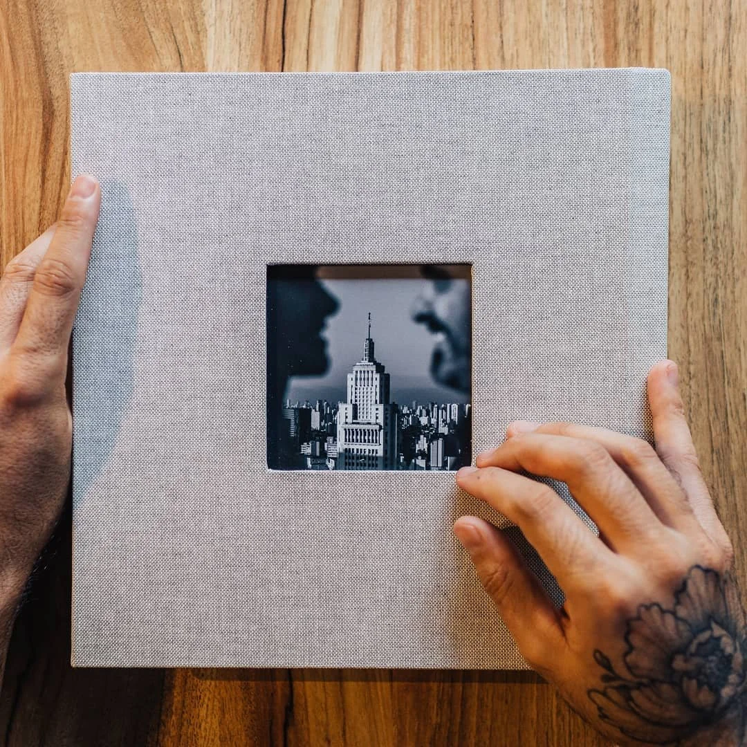 Mãos segurando uma moldura de fotos branca, com uma foto preto e branco de um arranha-céu e duas pessoas em perfil na frente.