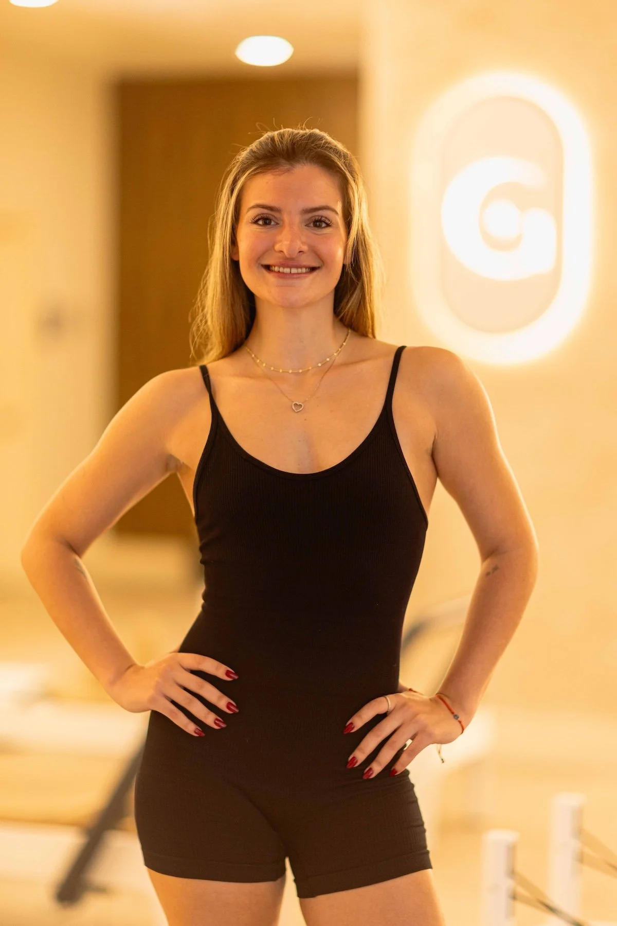 A young woman with long blonde hair, wearing a black tank top and shorts, smiling and standing with her hands on her hips indoors, with a bright light and logo in the background.
