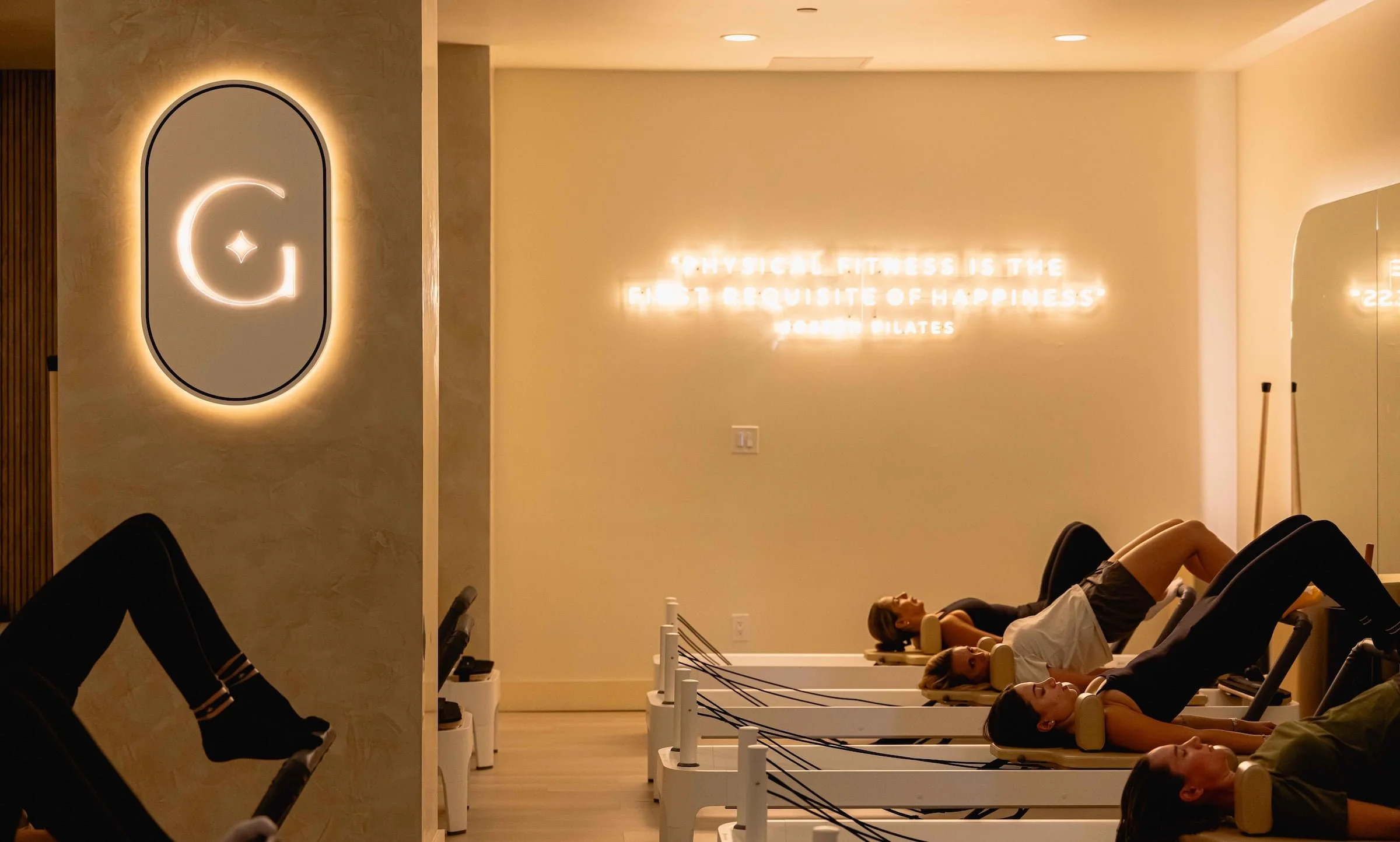 People lying on Pilates reformer machines in a fitness studio with soft lighting and motivational quotes on the wall.
