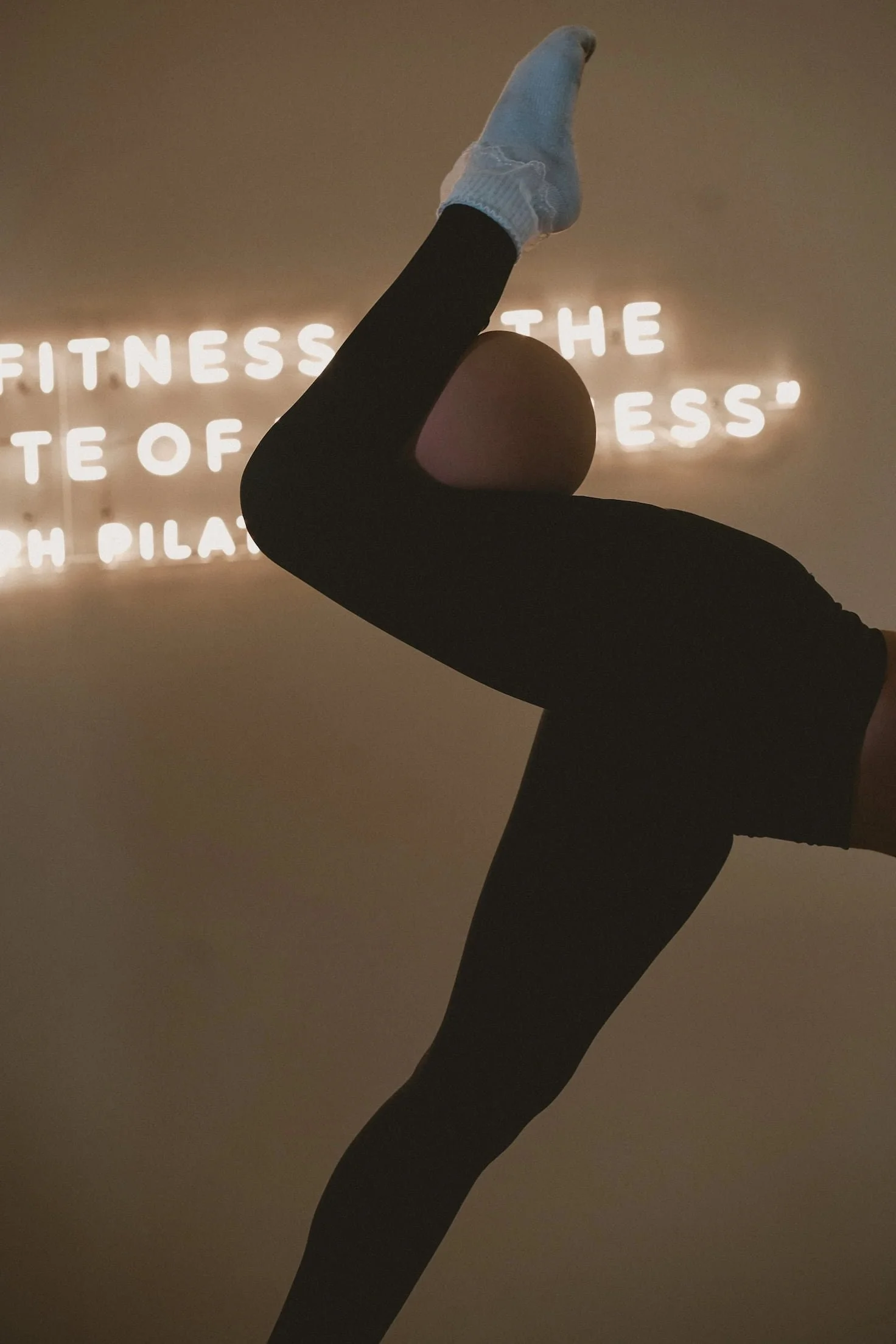 A person performing a backbend yoga pose, with one foot raised towards the ceiling, in front of a wall with illuminated text.