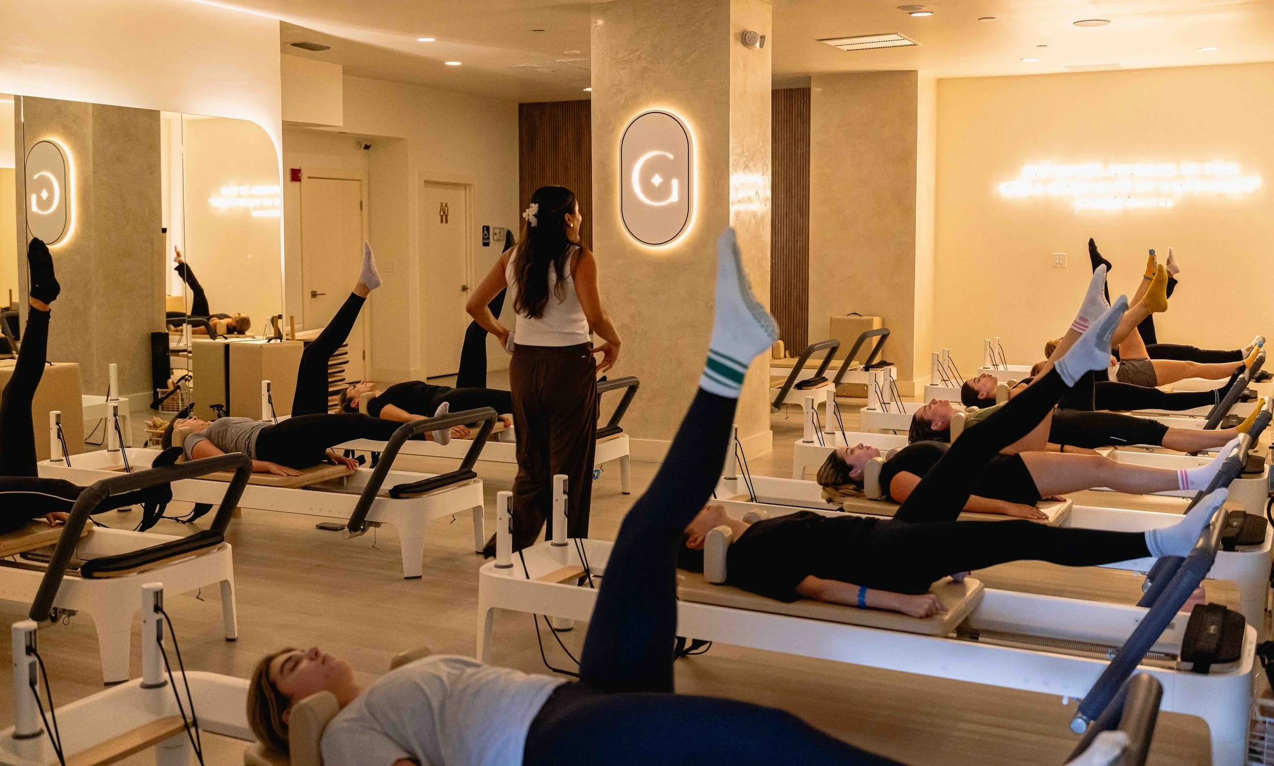 Group of women participating in a Pilates class at a fitness studio, lying on reformer machines with legs raised while an instructor oversees the session inside a modern, warmly lit room.