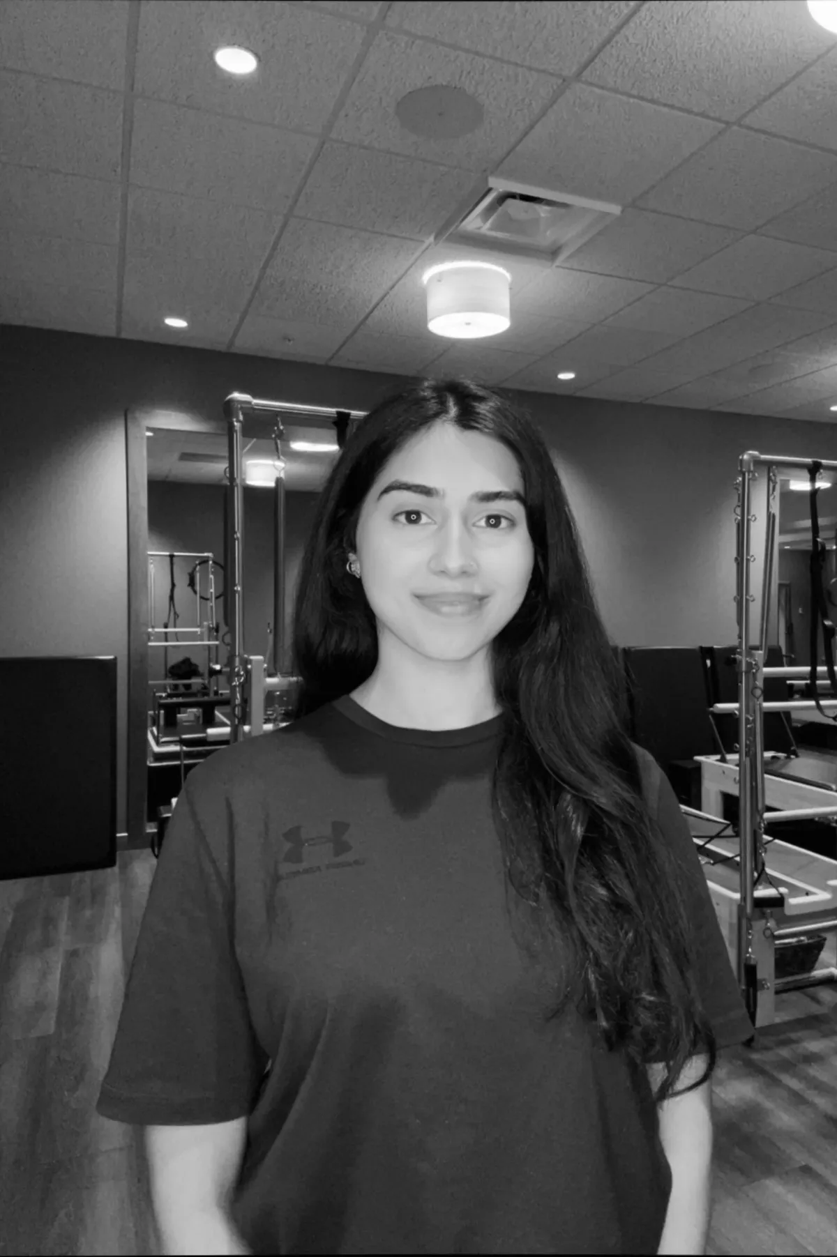 A woman with long dark hair and a smile standing in a fitness gym, wearing a dark sports shirt with the Under Armour logo, with workout equipment visible in the background.