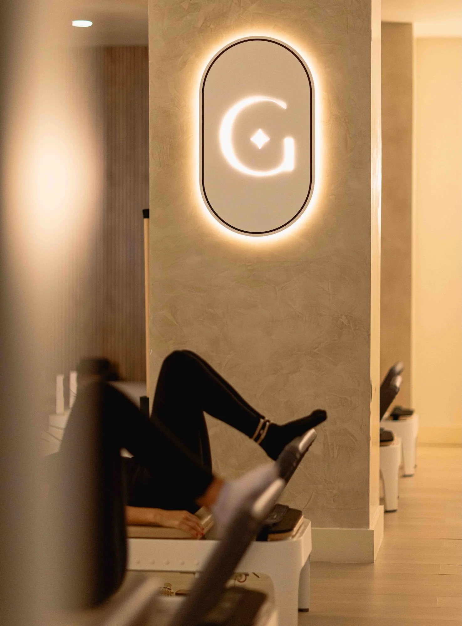 A person lying on an exercise bench with their legs up, in a gym or fitness studio. There is a glowing illuminated sign on the wall featuring a stylized logo.