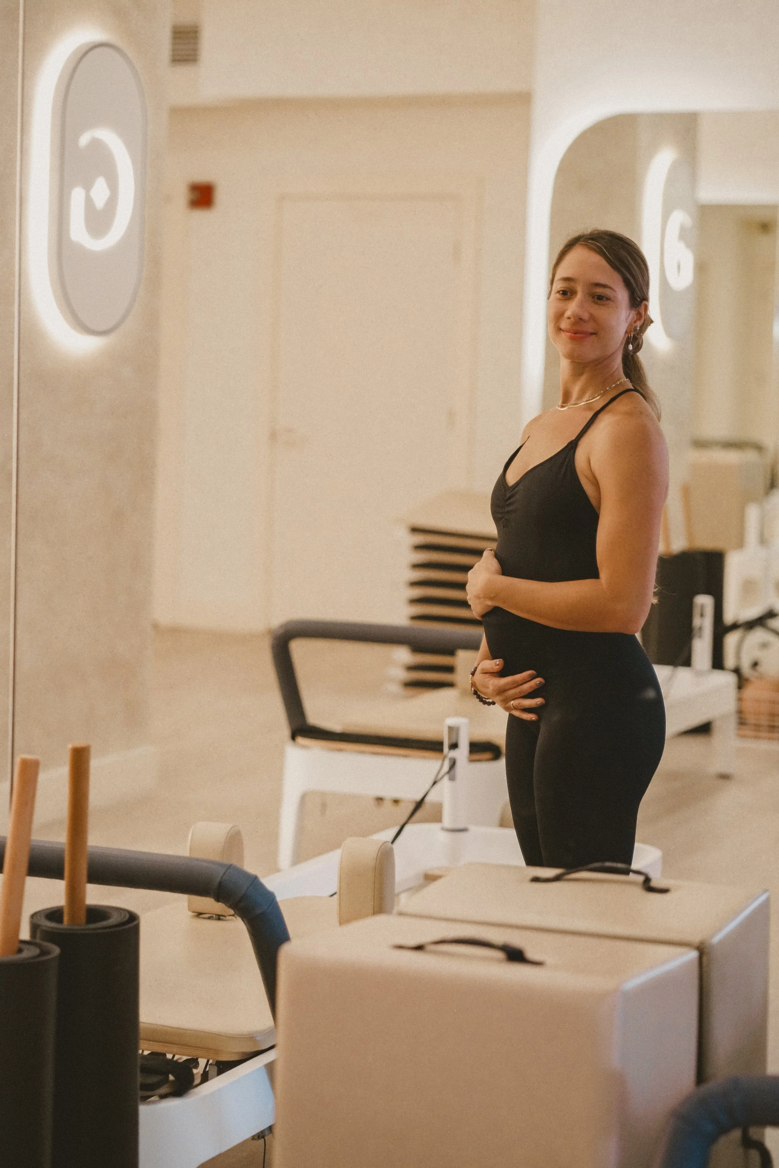 A woman standing in a fitness or wellness facility, smiling, with hand on her stomach, wearing a black workout outfit.