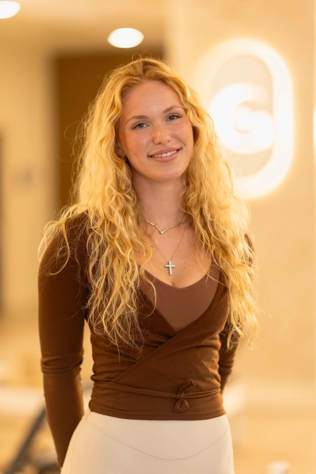 A young woman with long, curly blonde hair smiling in an indoor setting with warm lighting and the letter 'G' illuminated in the background.