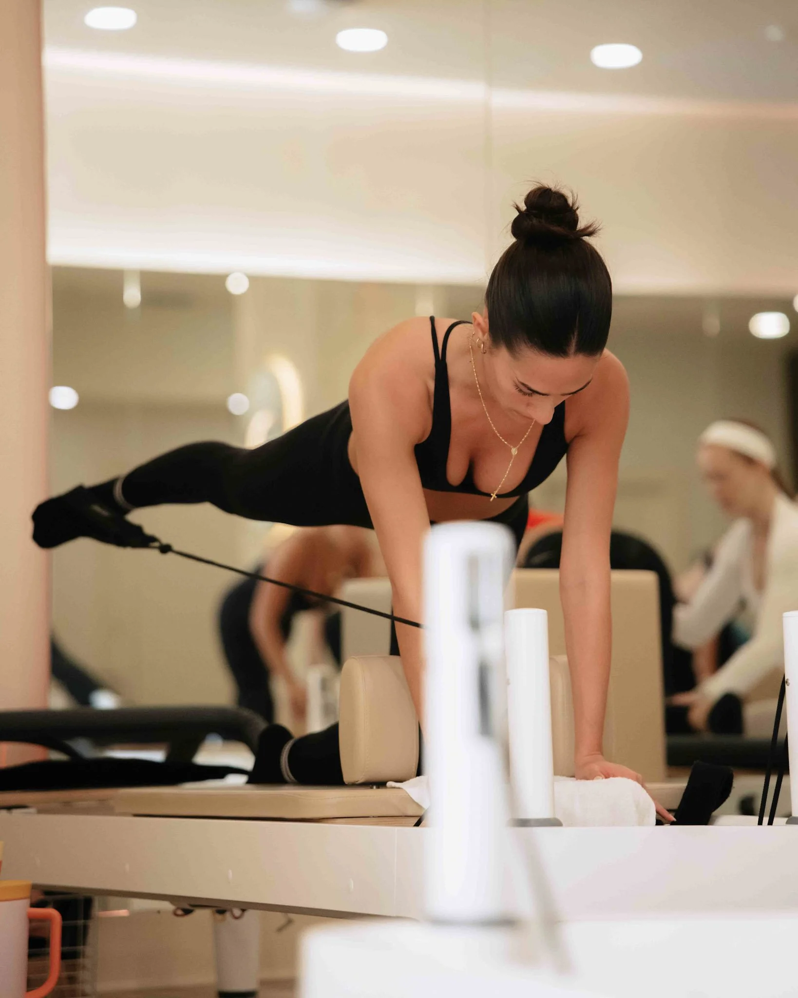 Woman in black workout attire performing a Pilates exercise on a reformer machine, with others exercising in the background.