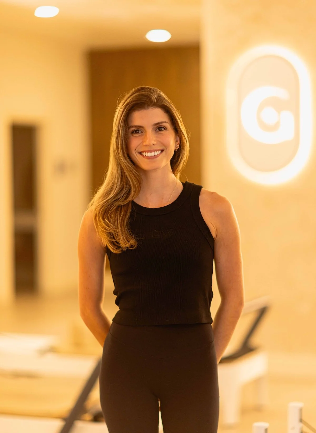 A smiling woman with long wavy hair in a black sleeveless top standing in a warmly lit indoor space.