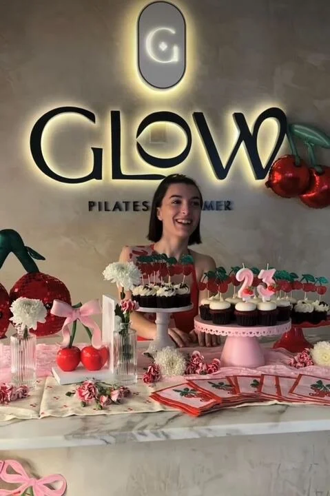 A girl at a birthday celebration with a table of cupcakes and desserts, decorated with cherries and flowers, in front of a wall with a GLOW logo. The setting appears to be in a Pilates studio.