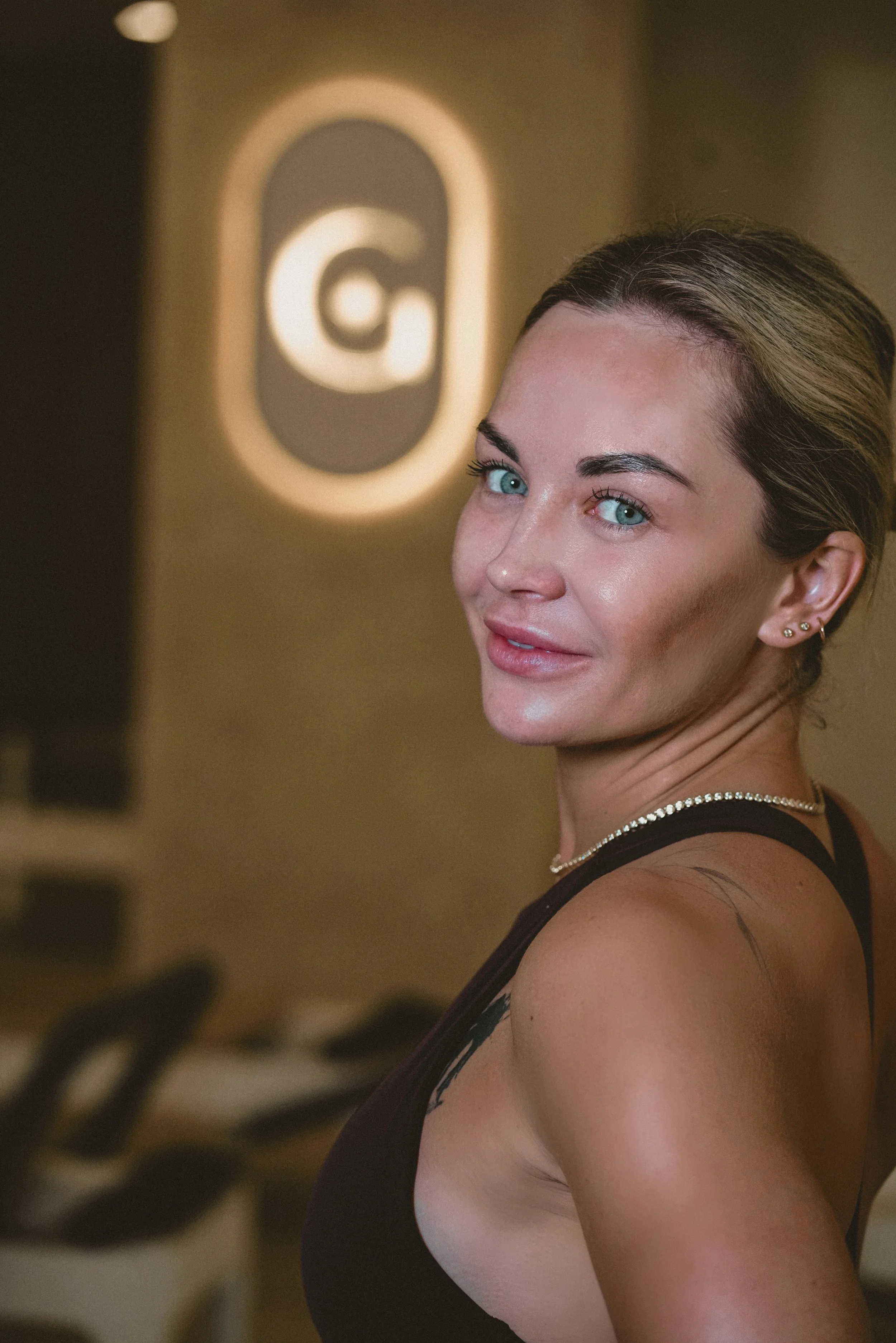 A woman with blonde hair and blue eyes smiling at the camera, standing in front of a blurred background with a logo behind her.