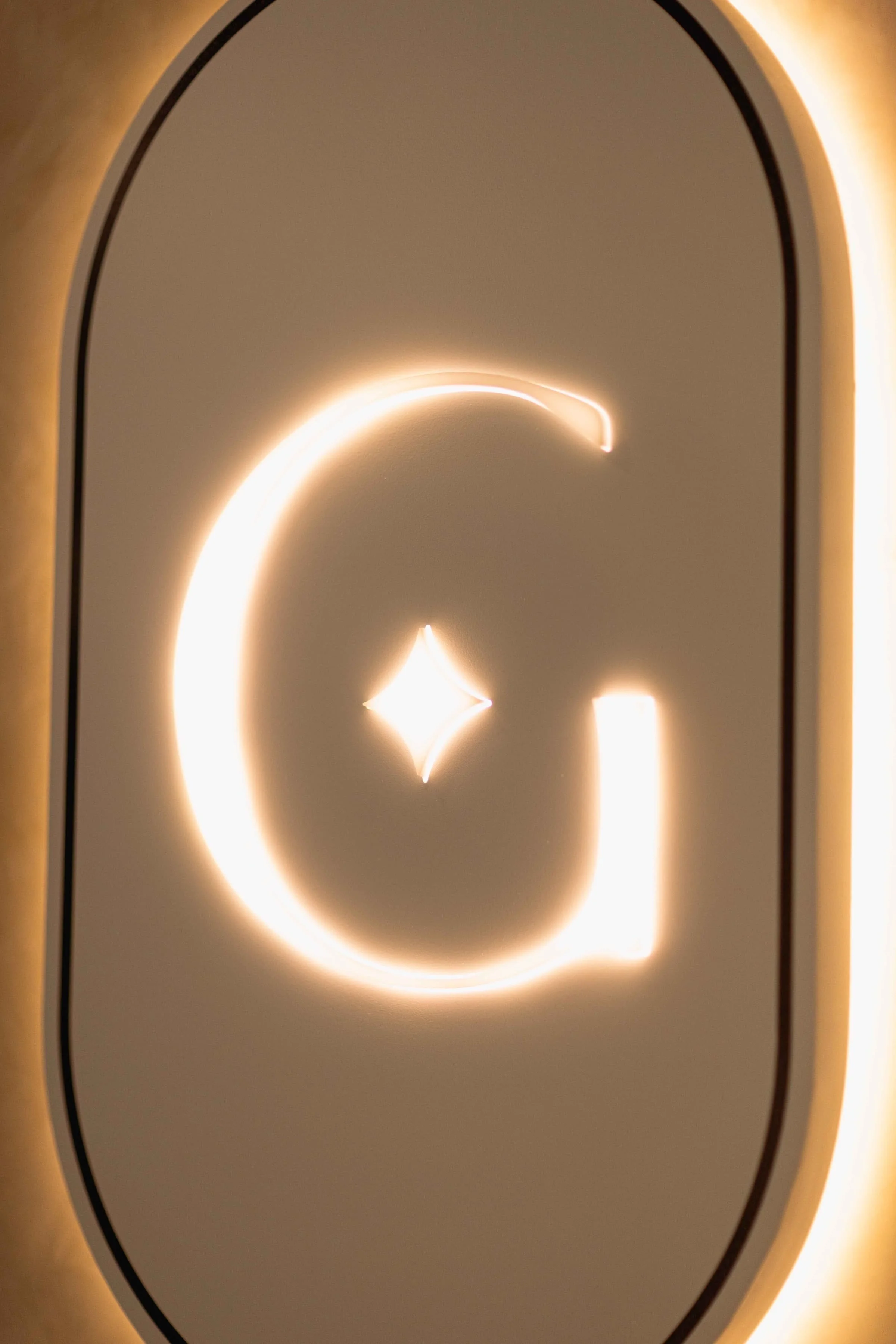 Neon sign with a large letter G and a small diamond shape in the center, illuminated against a light-colored background.