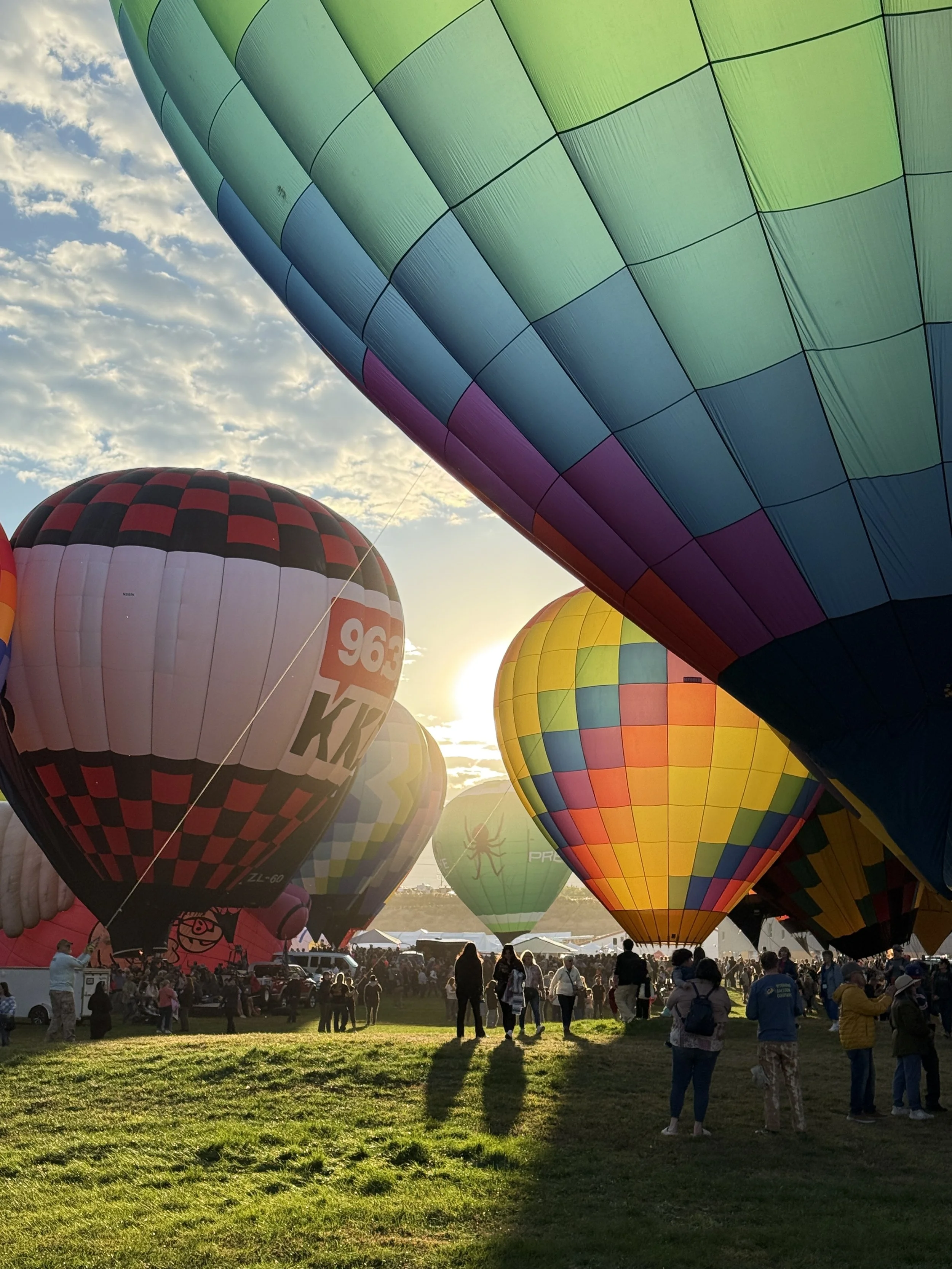 Hot air balloons preparing for takeoff at sunrise during a festival, with people walking and gathered on a grassy field.